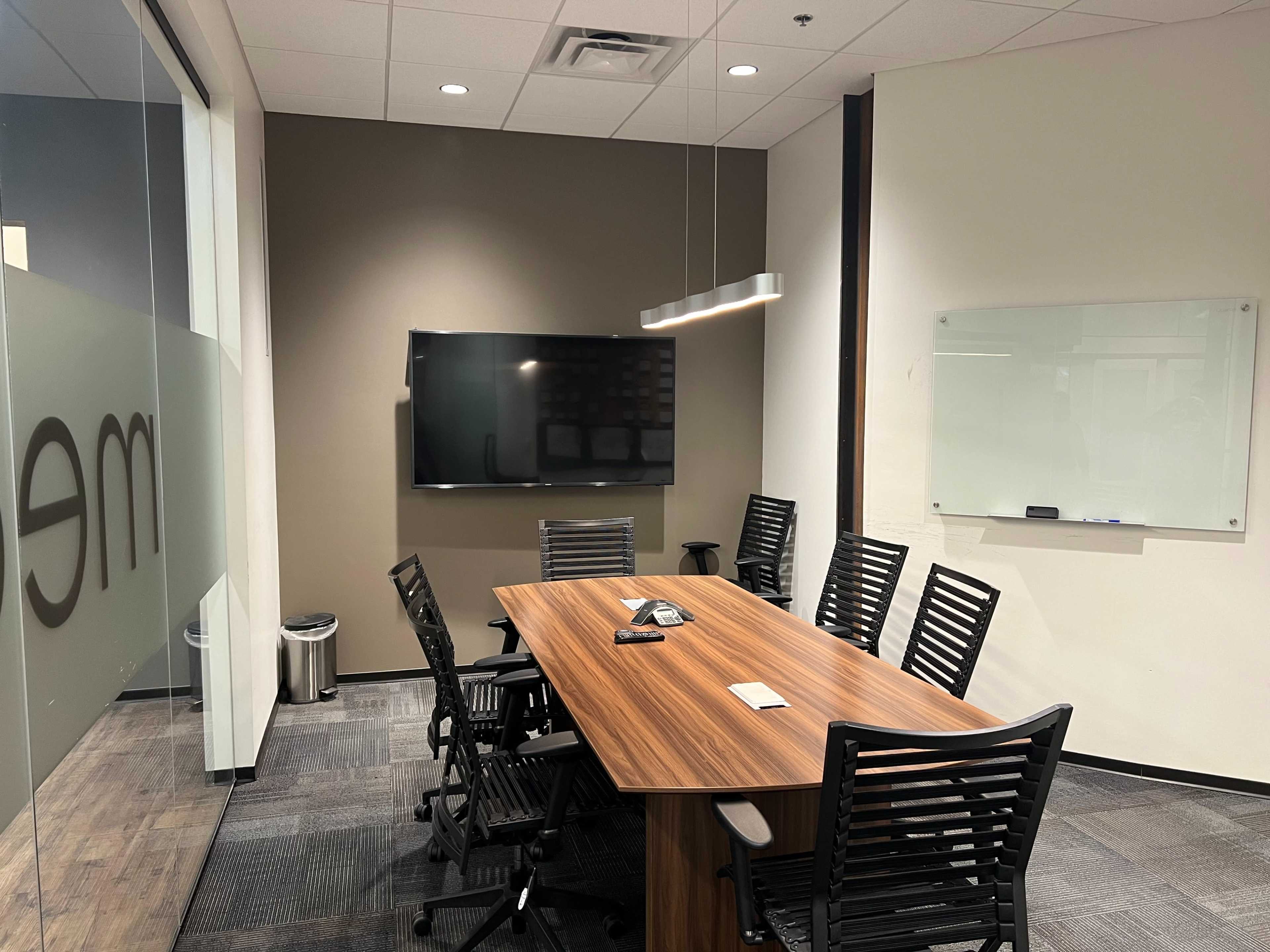 The image shows a conference room featuring a large wooden table surrounded by black chairs, a wall-mounted television, and a whiteboard.