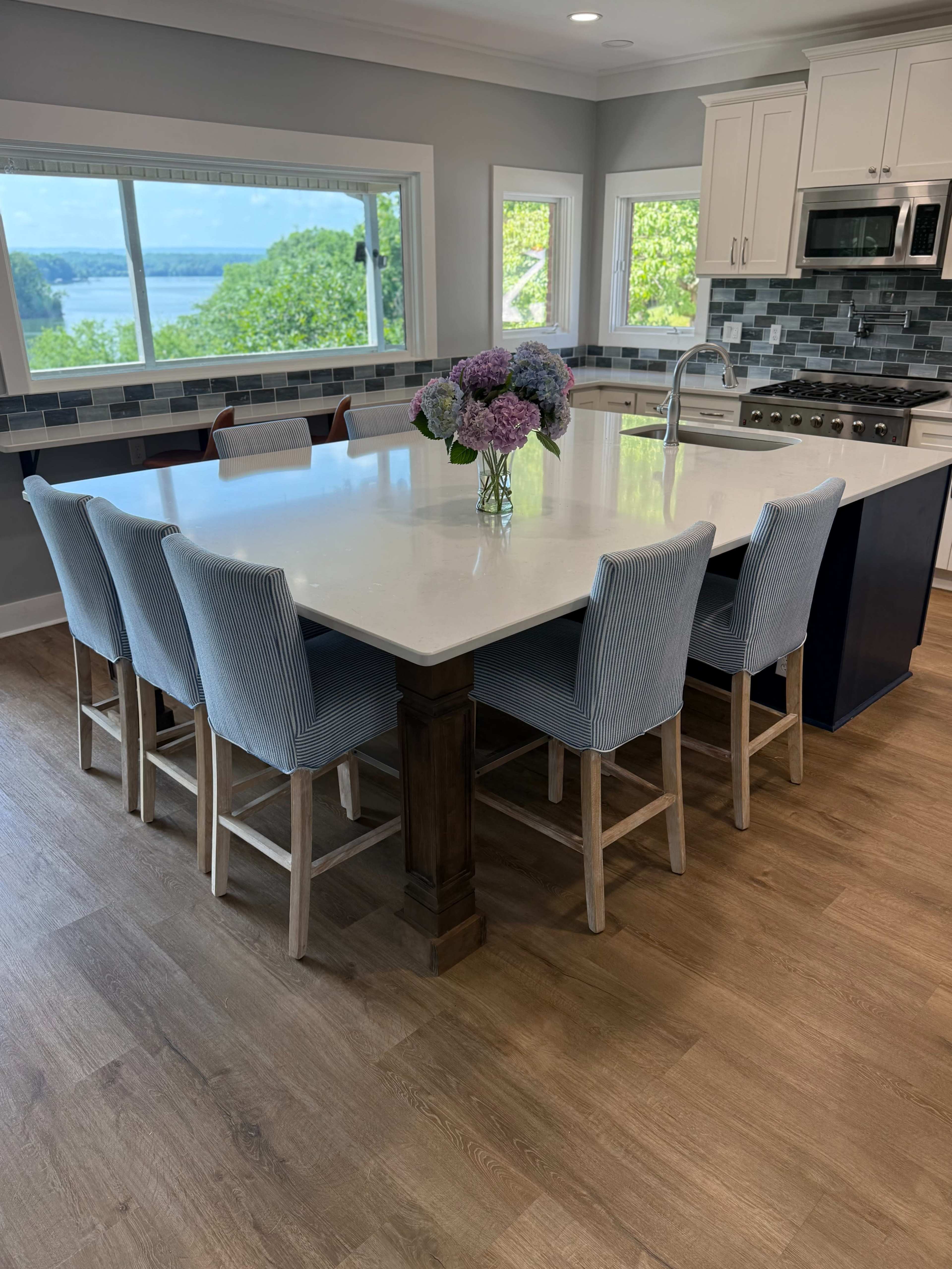 A modern kitchen features a large island with seating for six, surrounded by light blue upholstered bar stools and a view of the water through large windows.
