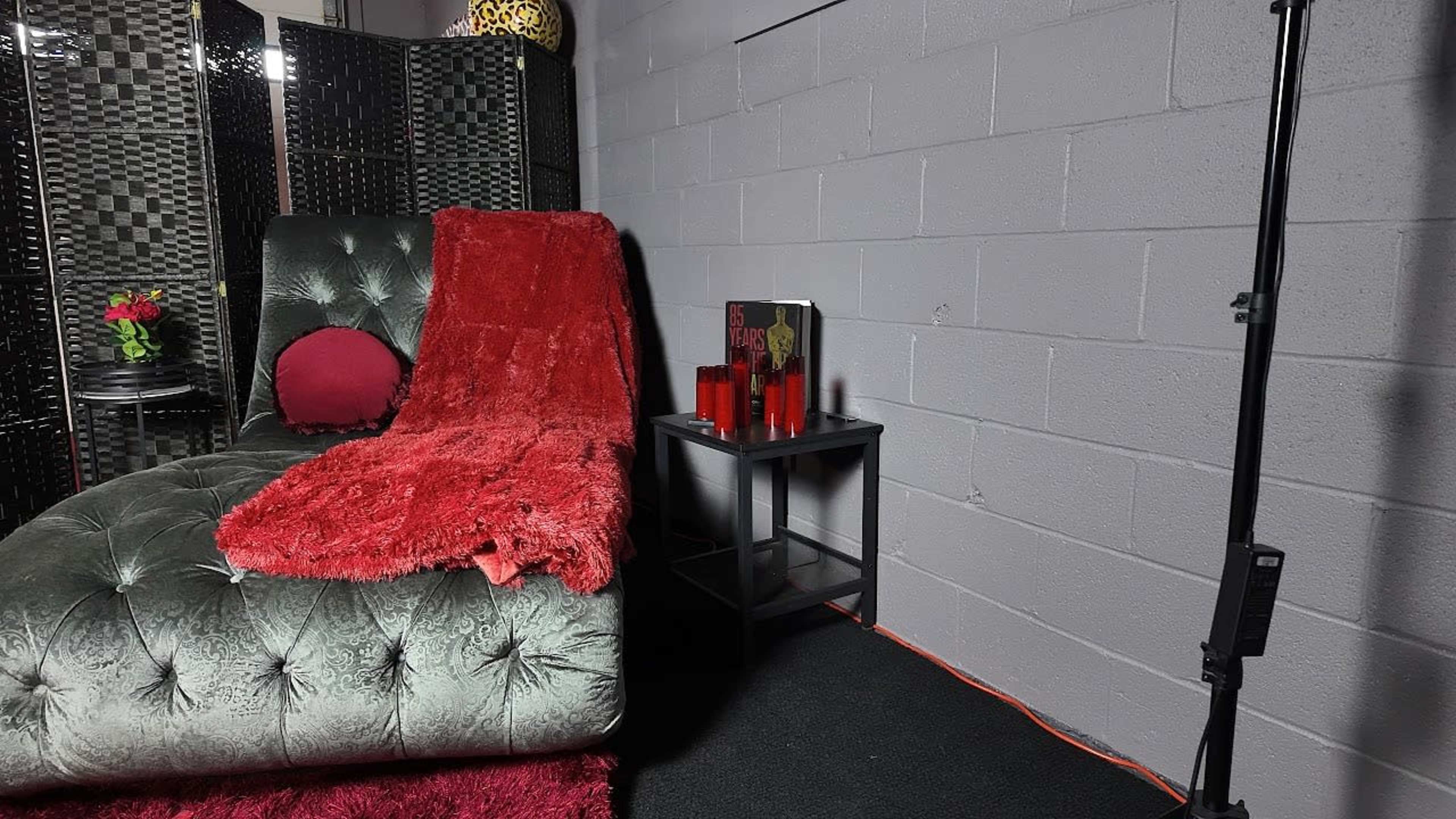 The image shows a cozy indoor setting with a green tufted chaise lounge draped with a red throw blanket, accompanied by a small black side table holding a stack of books and red candles.