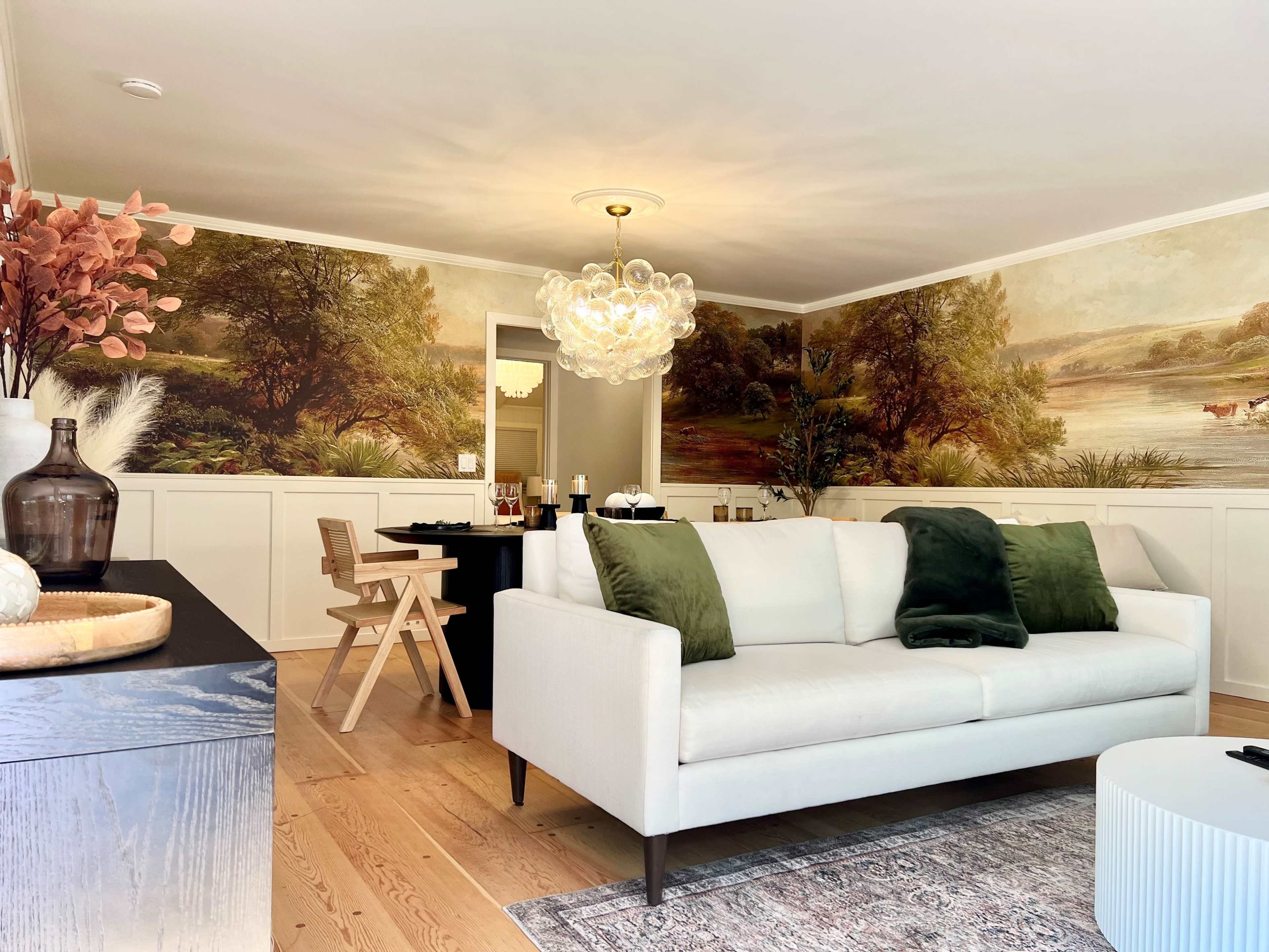 A modern living room with a white sofa, a wooden dining table, and a scenic mural depicting nature on the walls.