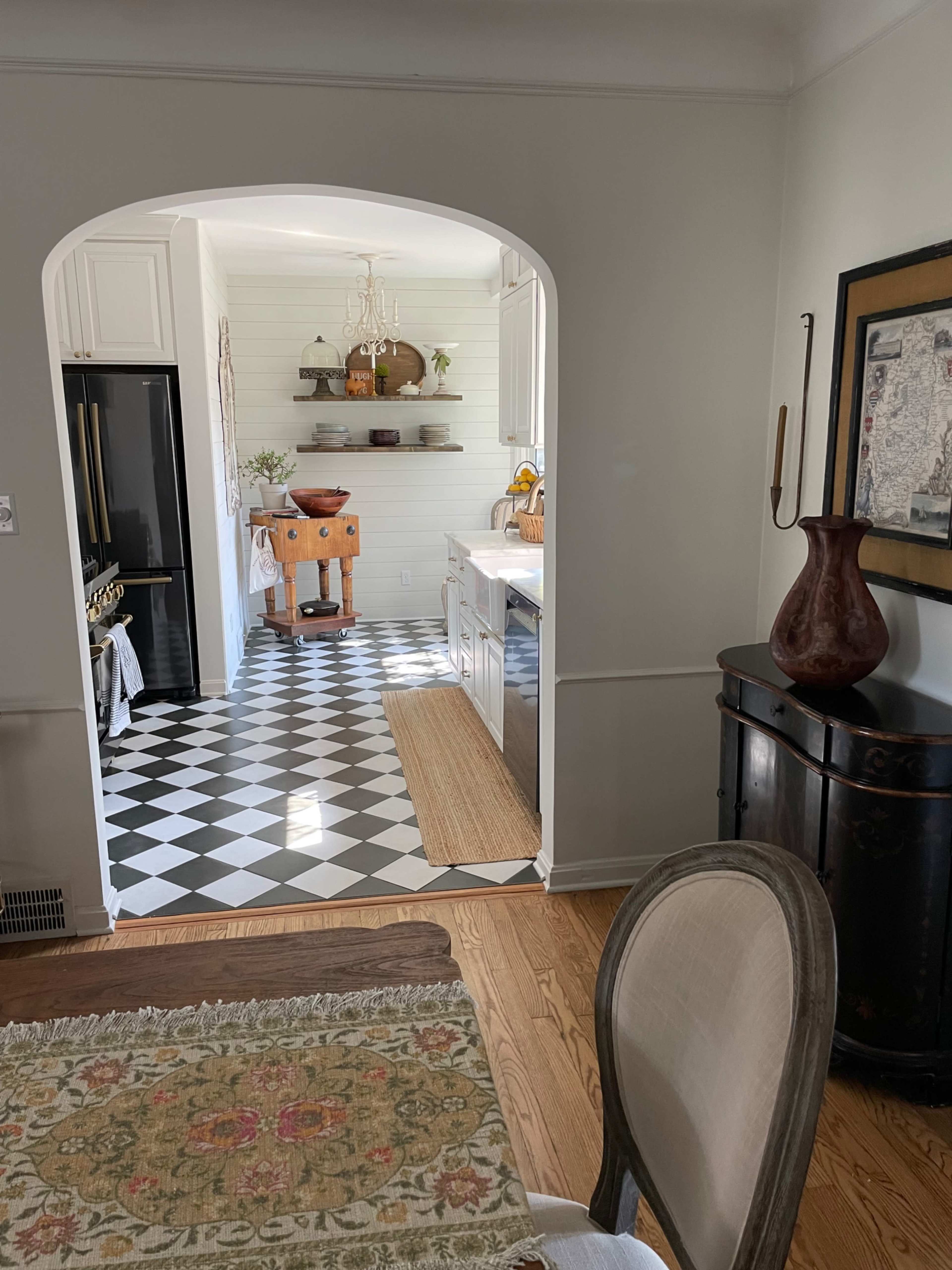 The image shows a kitchen area visible through an arched doorway, featuring a checkered black and white floor and open shelving with decorative items.