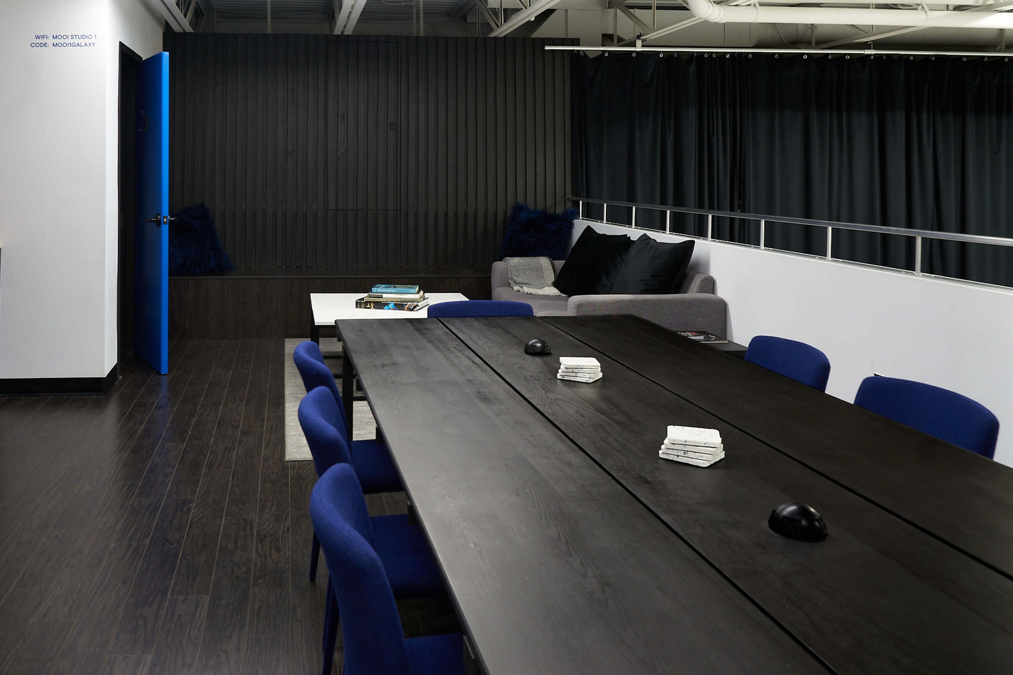 A long black conference table with blue chairs is positioned in a modern meeting room featuring a gray sofa and a dark wall with curtains.
