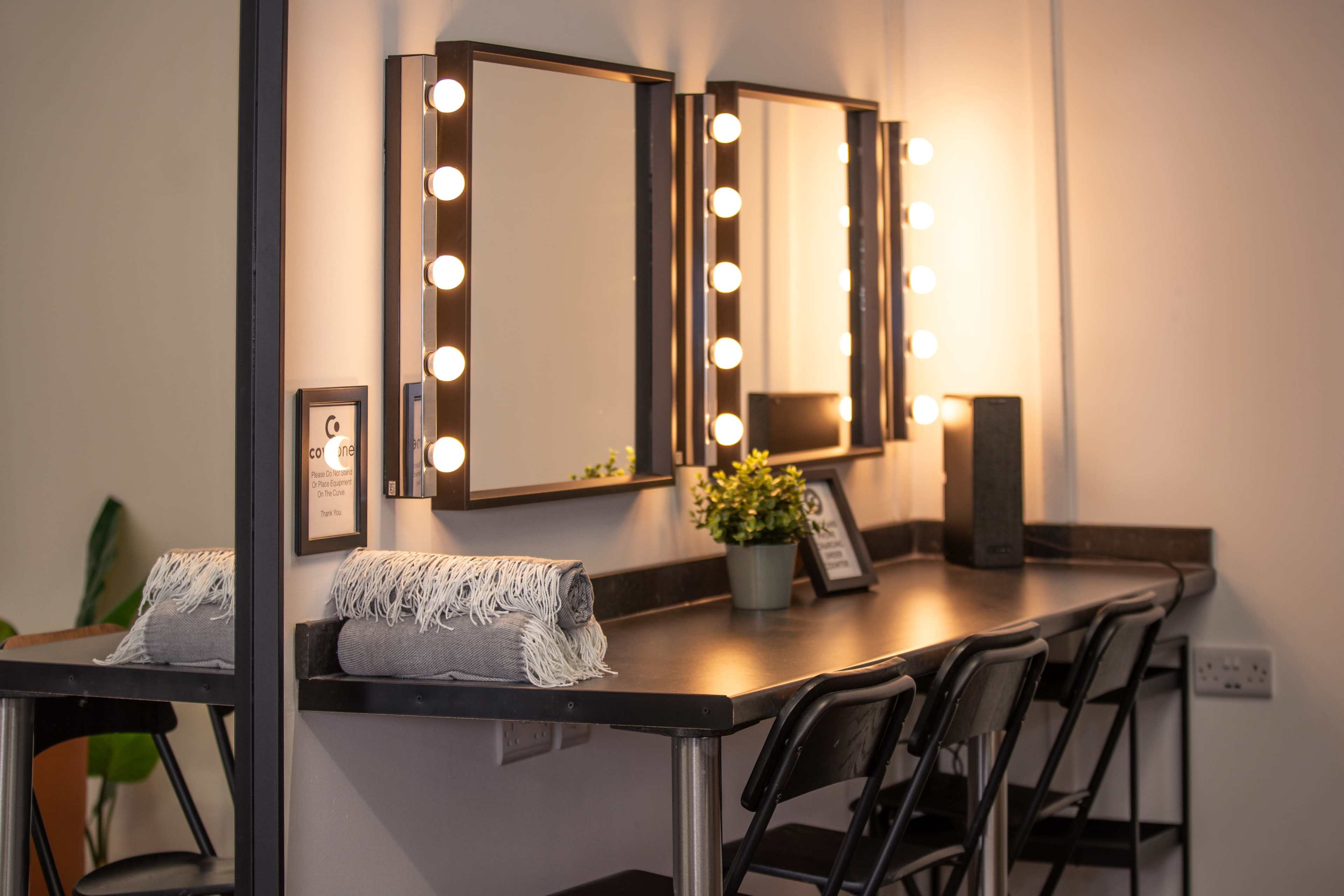 The image shows a makeup vanity with two lighted mirrors, a small potted plant, and several chairs arranged around a black countertop.