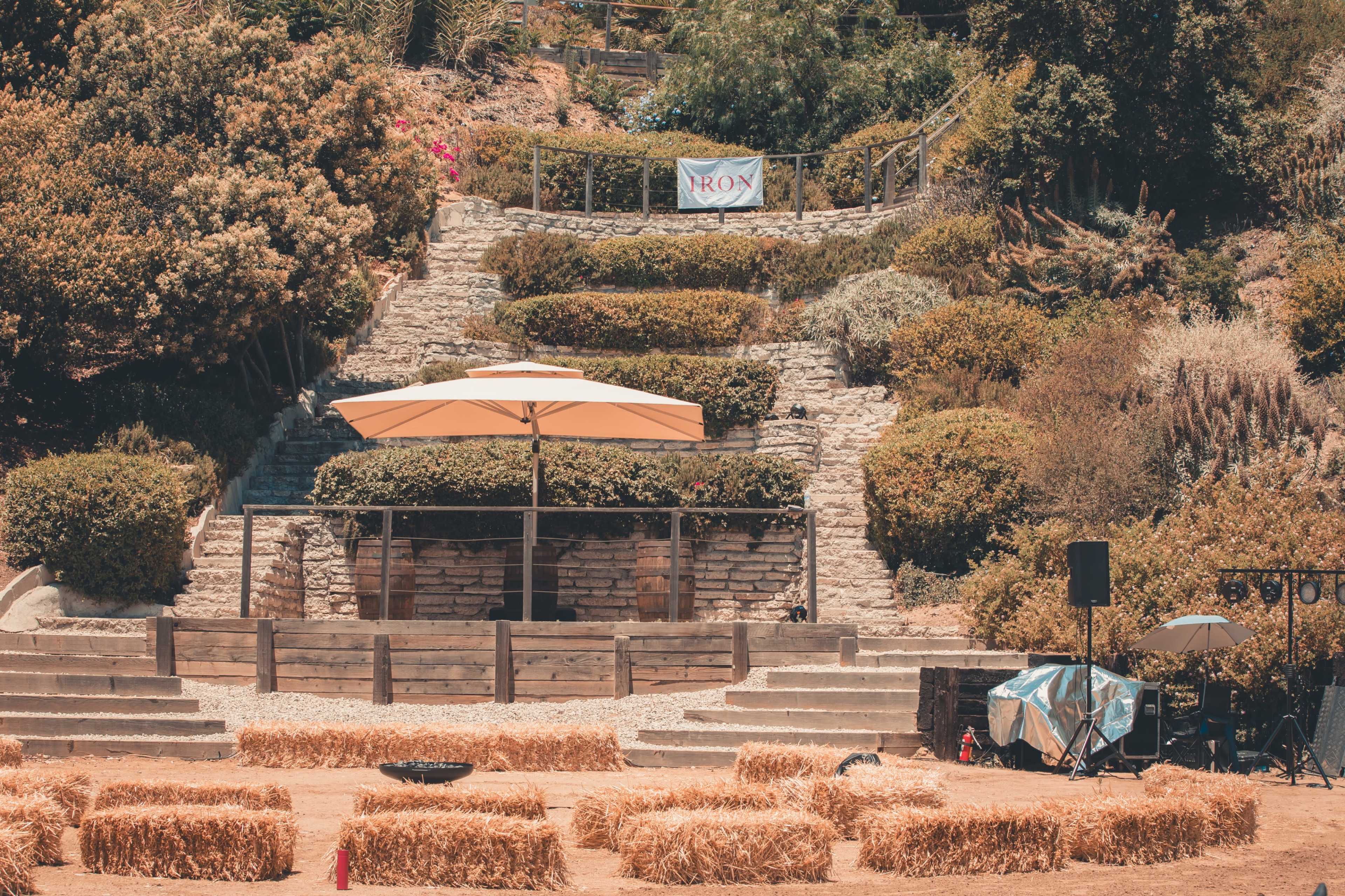 The image shows a terraced outdoor area with a banner, an umbrella over a small stage, and hay bales arranged in front.
