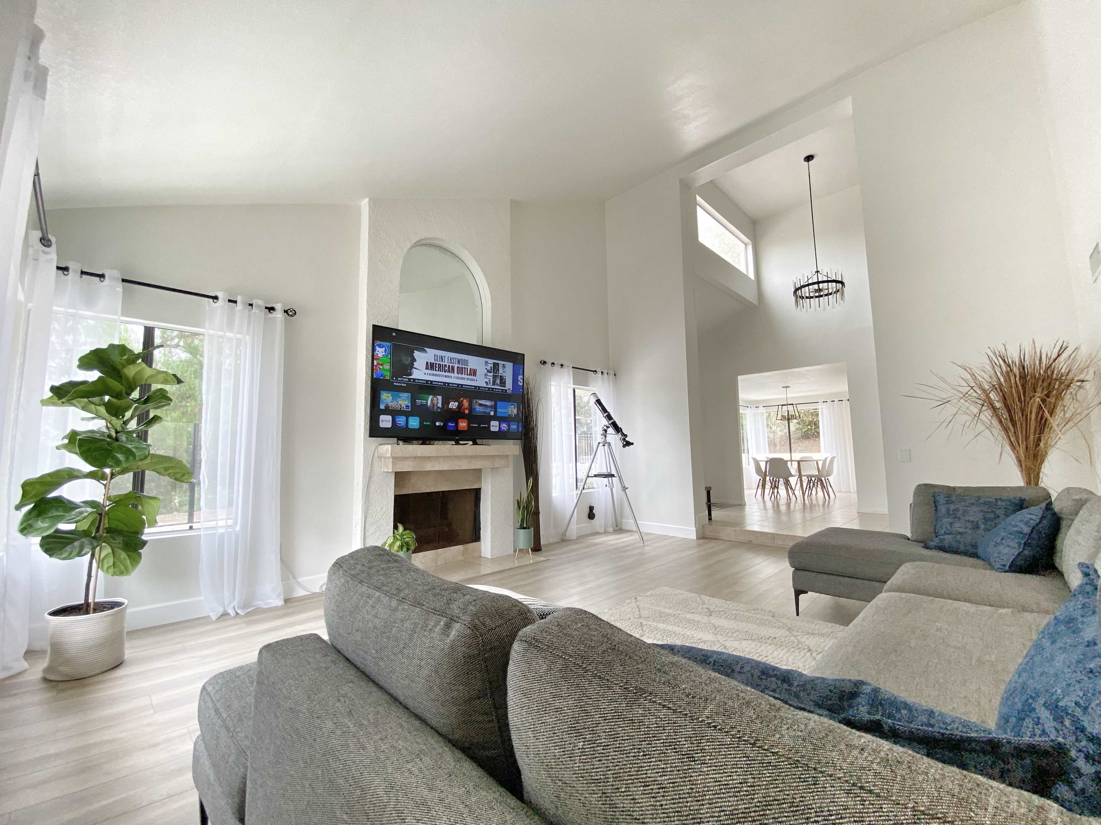 A modern living room with a gray sectional sofa, a large wall-mounted television, a potted plant, and a telescope beside a window.