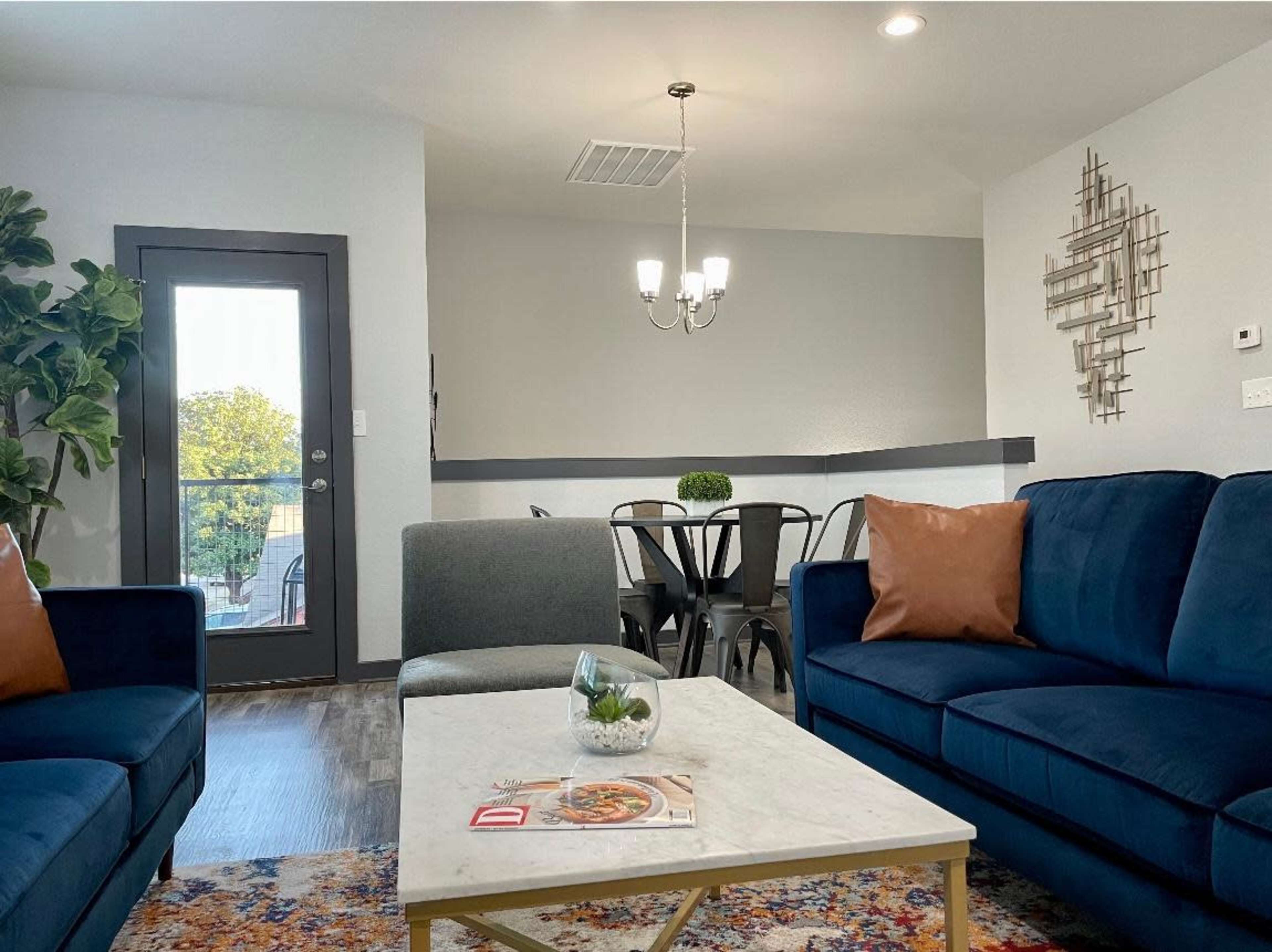 A modern living room features two blue sofas, a marble coffee table, and a dining area with metal chairs, illuminated by a chandelier.