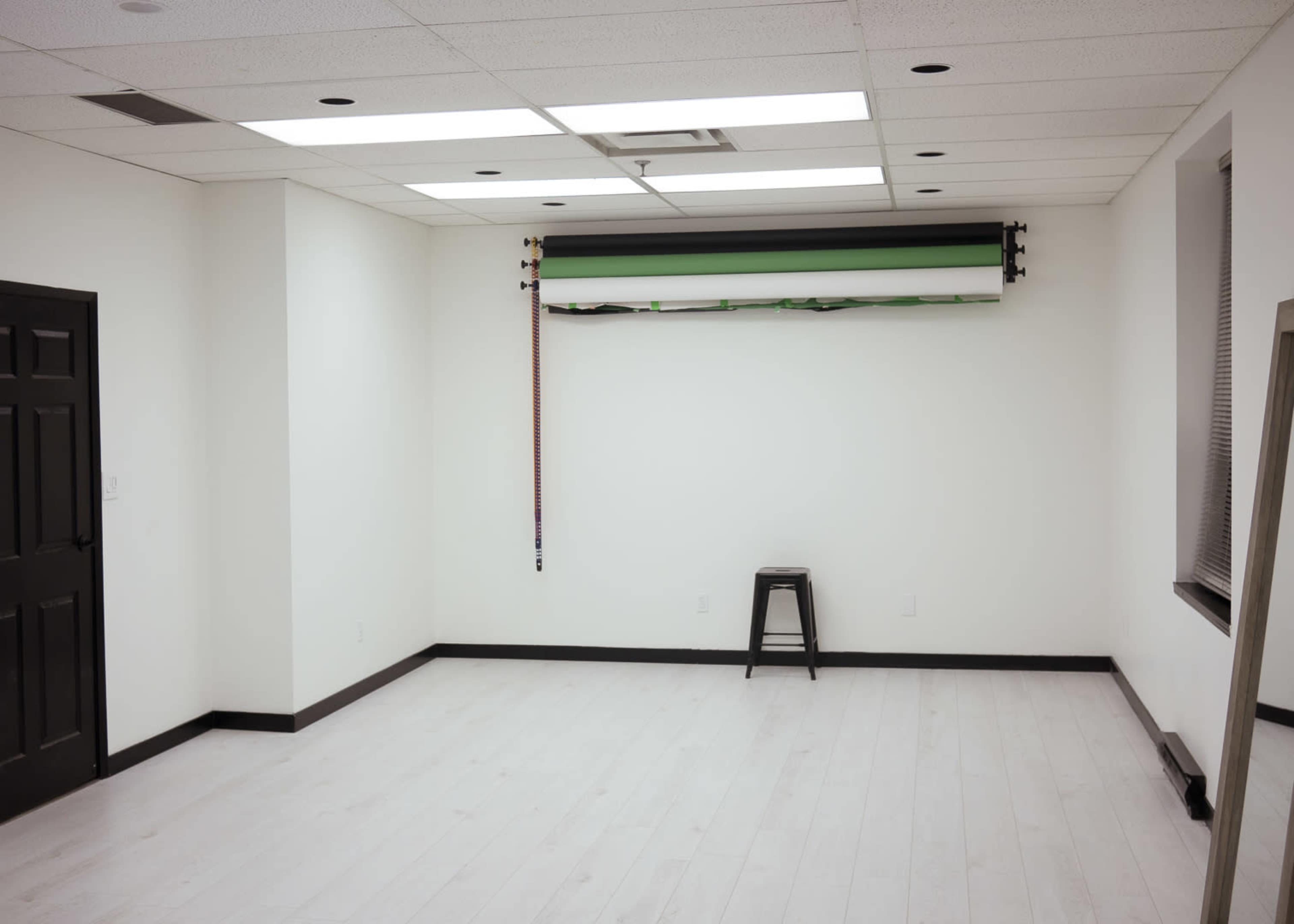 An empty room with white walls, a black door, and a single black stool in front of a rolled-up backdrop.