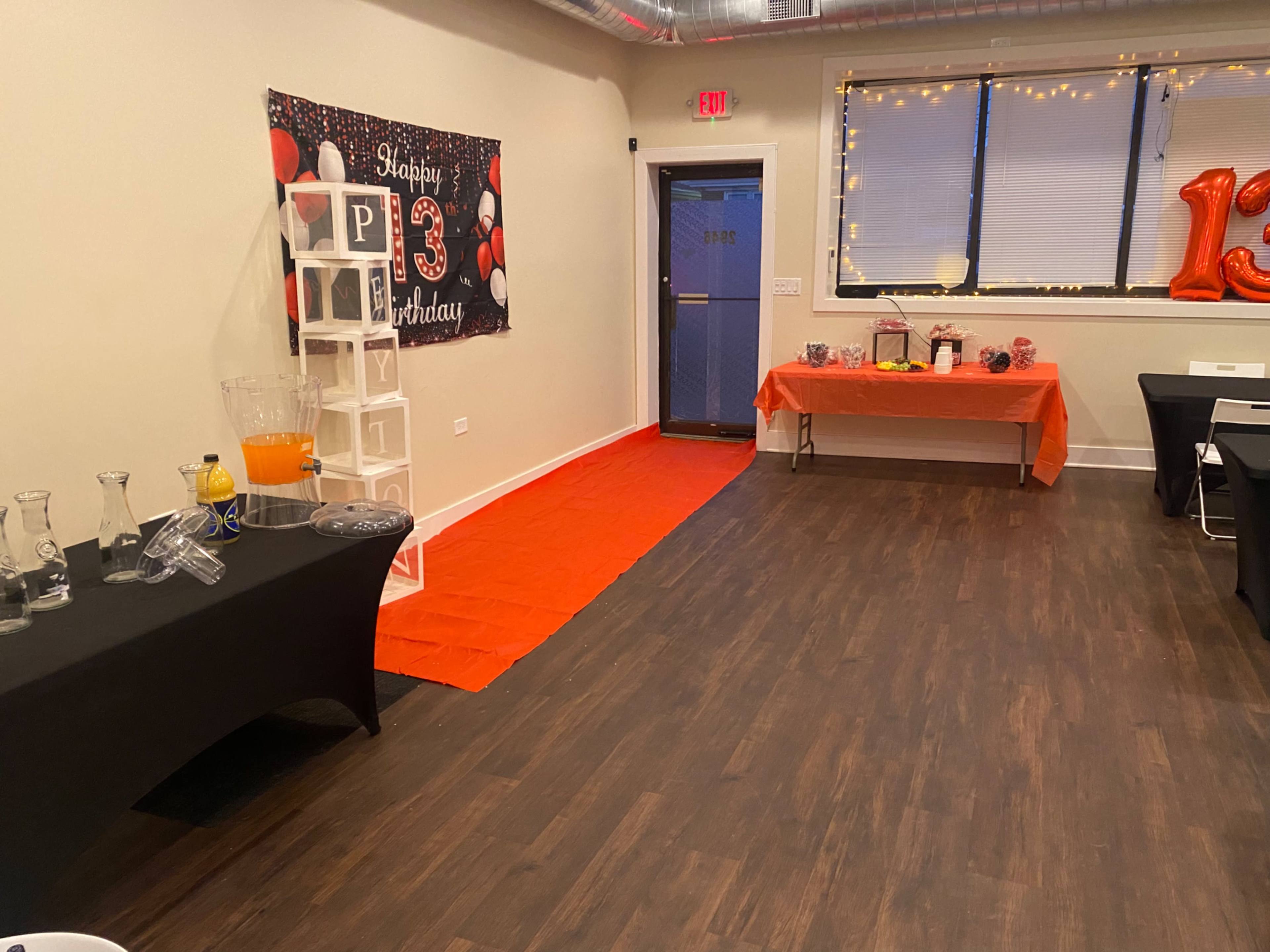 The interior of a party room features tables covered in black cloth, a decorative "Happy 13th Birthday" banner, and a bright orange carpet leading to a doorway.