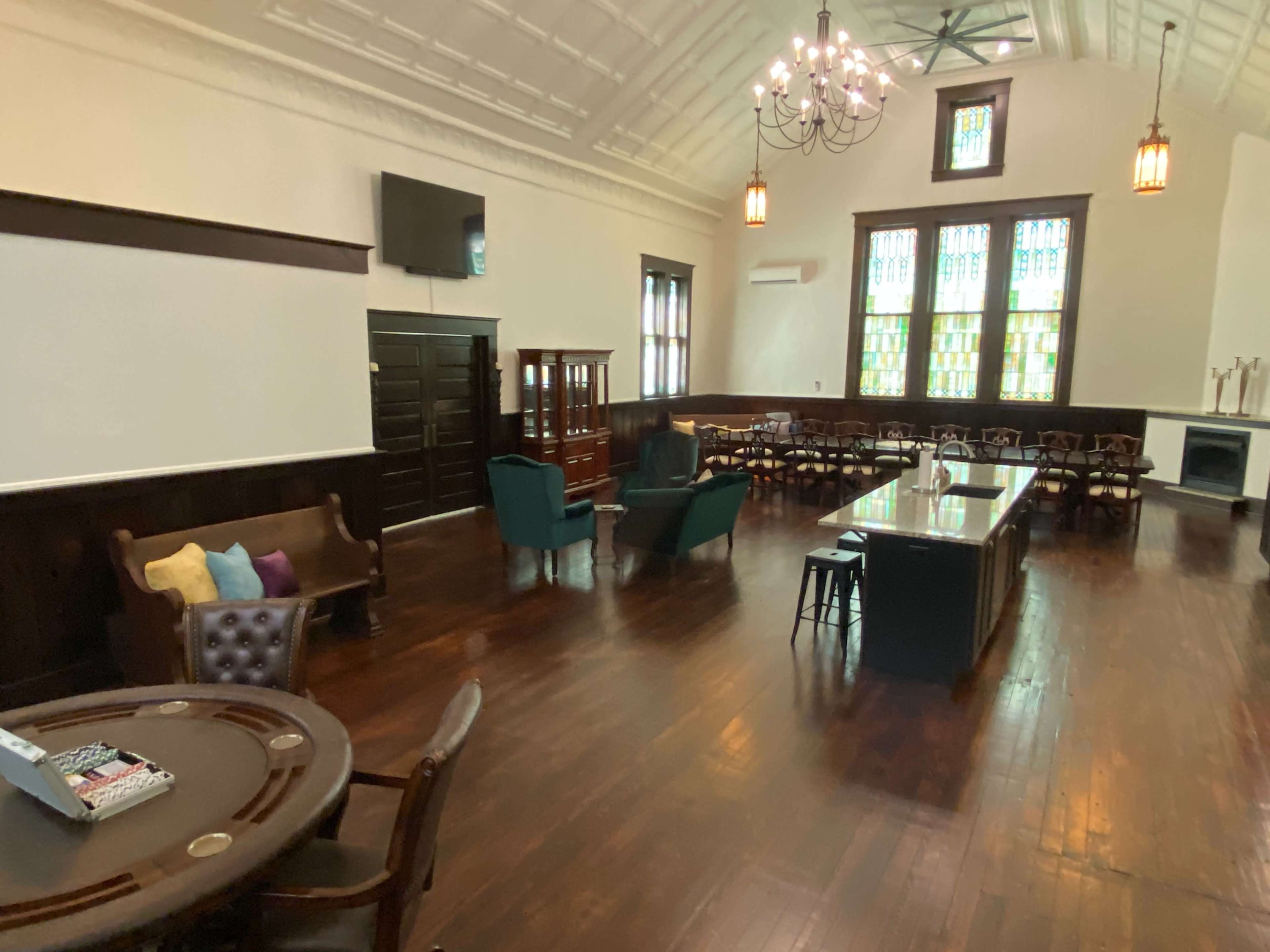 The image shows a spacious interior room with wooden floors, a large table, several seating areas, and stained glass windows.