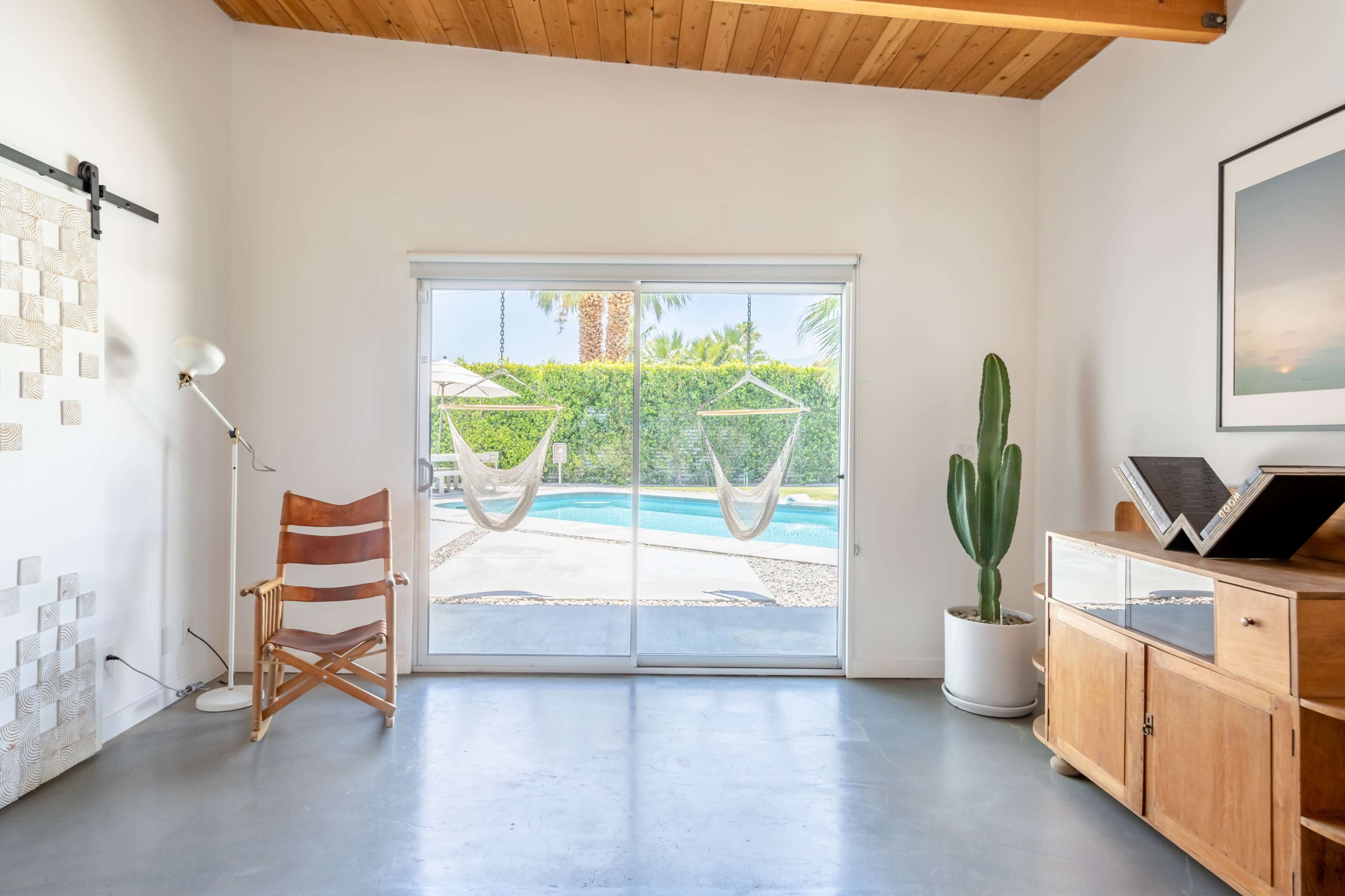 A bright room features a wooden ceiling, a large sliding glass door overlooking a pool, and minimalist furniture including a wooden chair and a potted cactus.