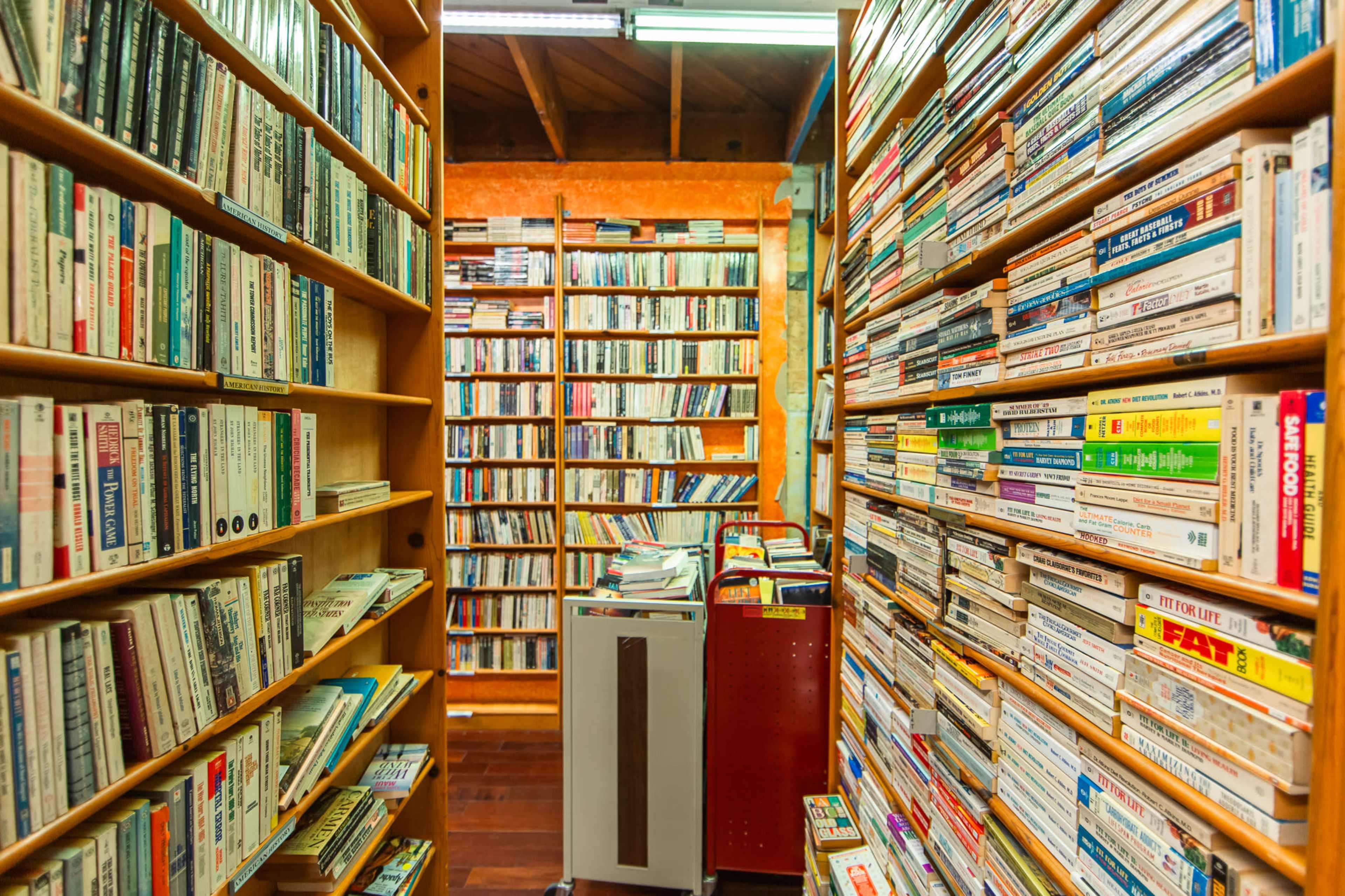 The image shows an aisle between two tall shelves filled with books and tapes in a cozy library or bookstore setting.