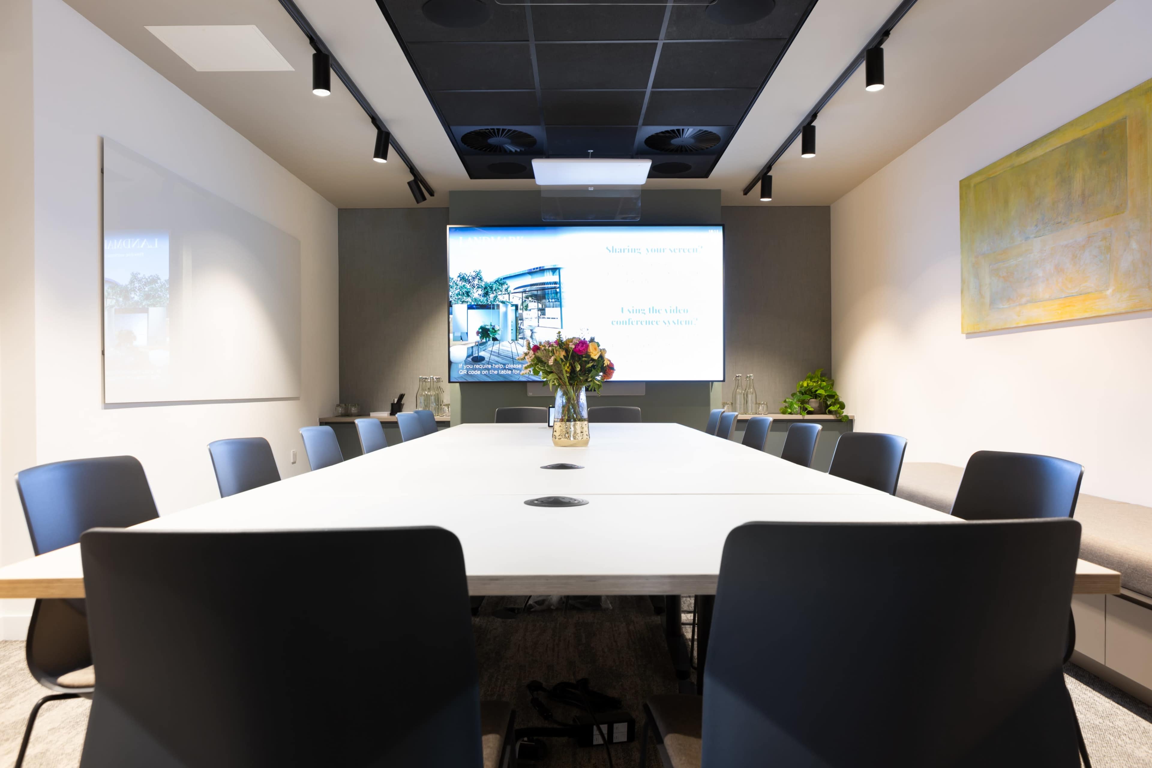 A modern conference room is set up with a long white table surrounded by black chairs, and a screen displaying a presentation at one end.