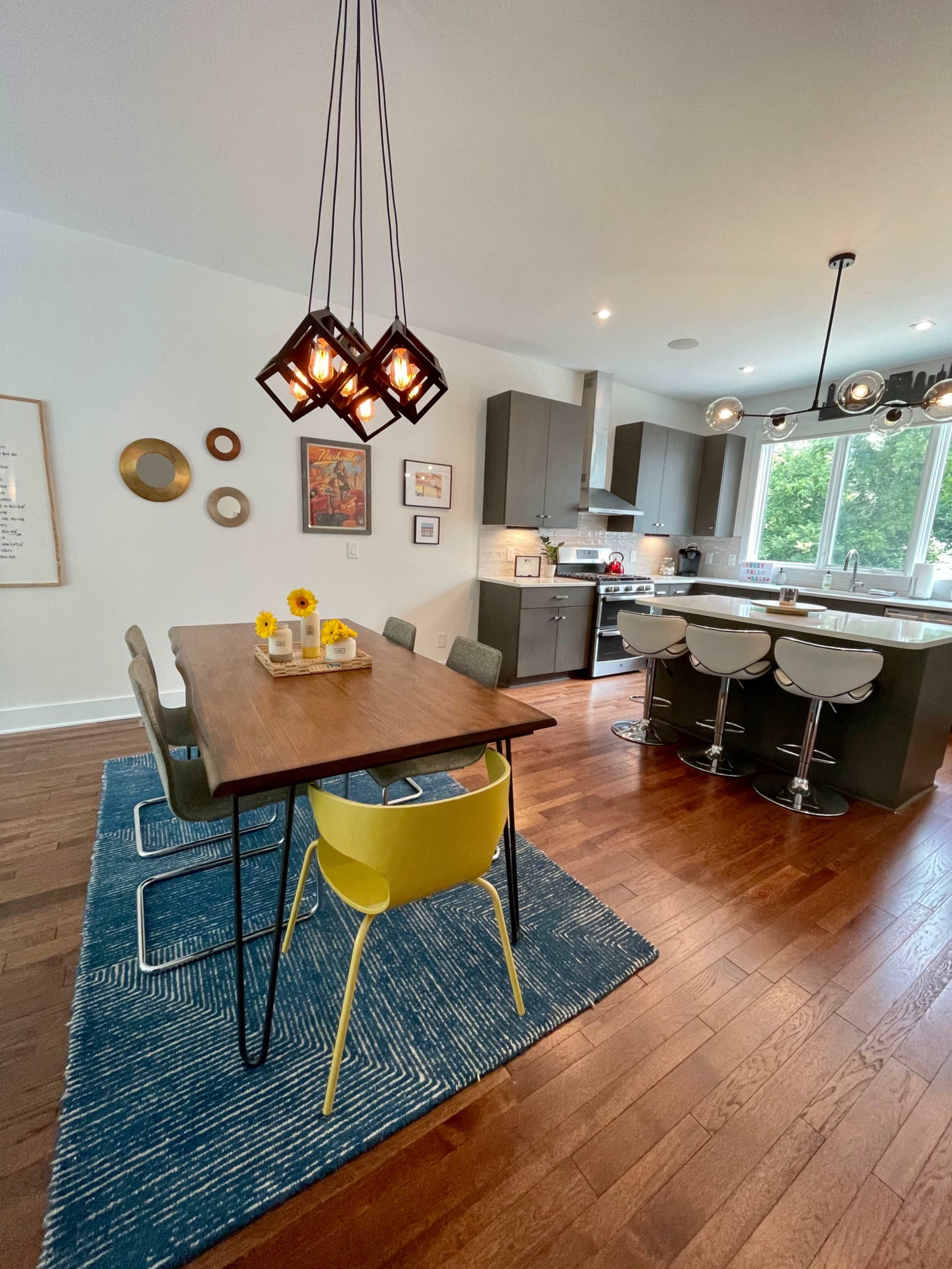 A modern kitchen and dining area features a wooden table surrounded by chairs, a blue rug, and pendant lighting.