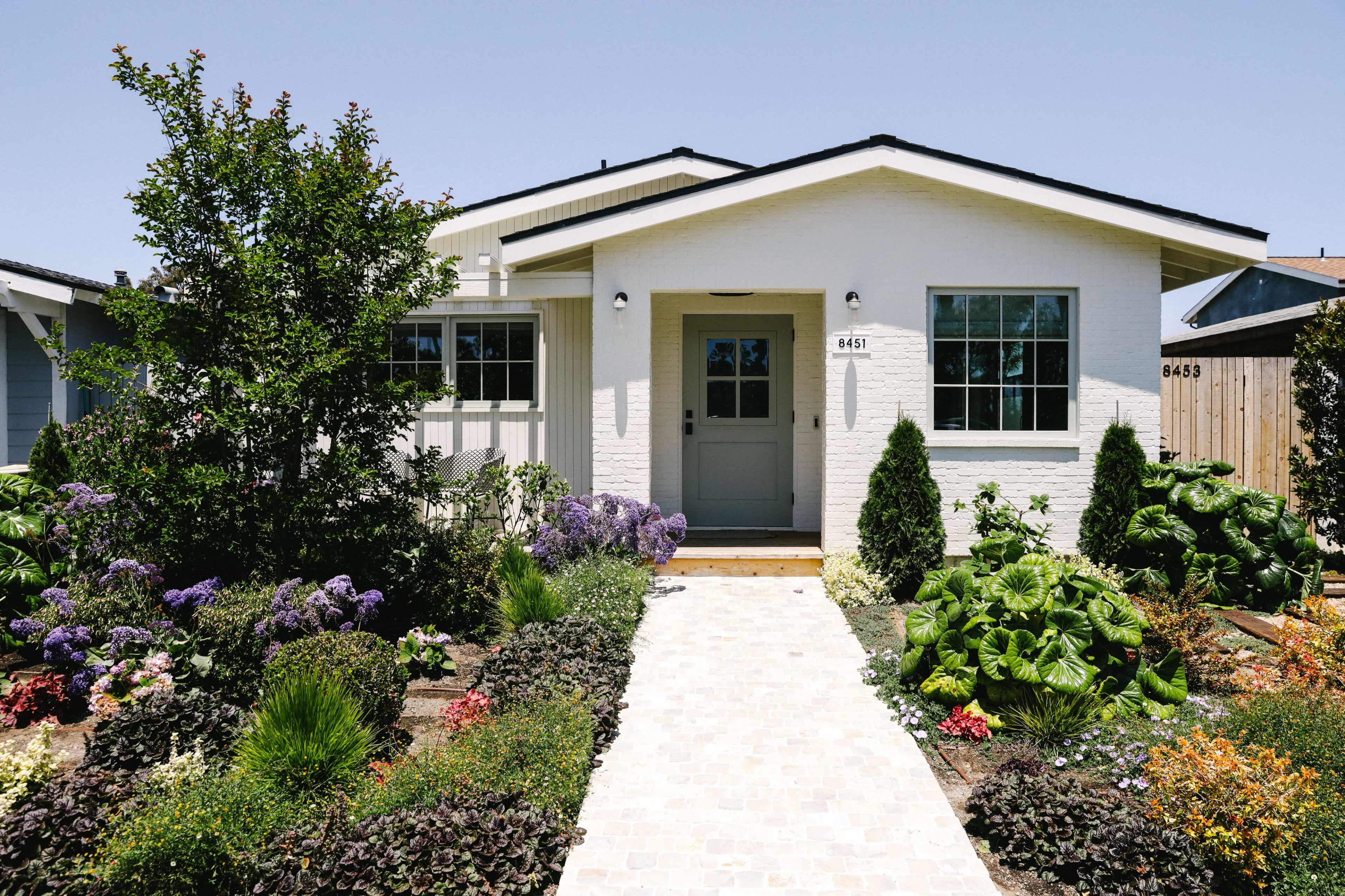 A modern white house with a front door flanked by shrubs and a well-maintained garden featuring various flowers and plants.
