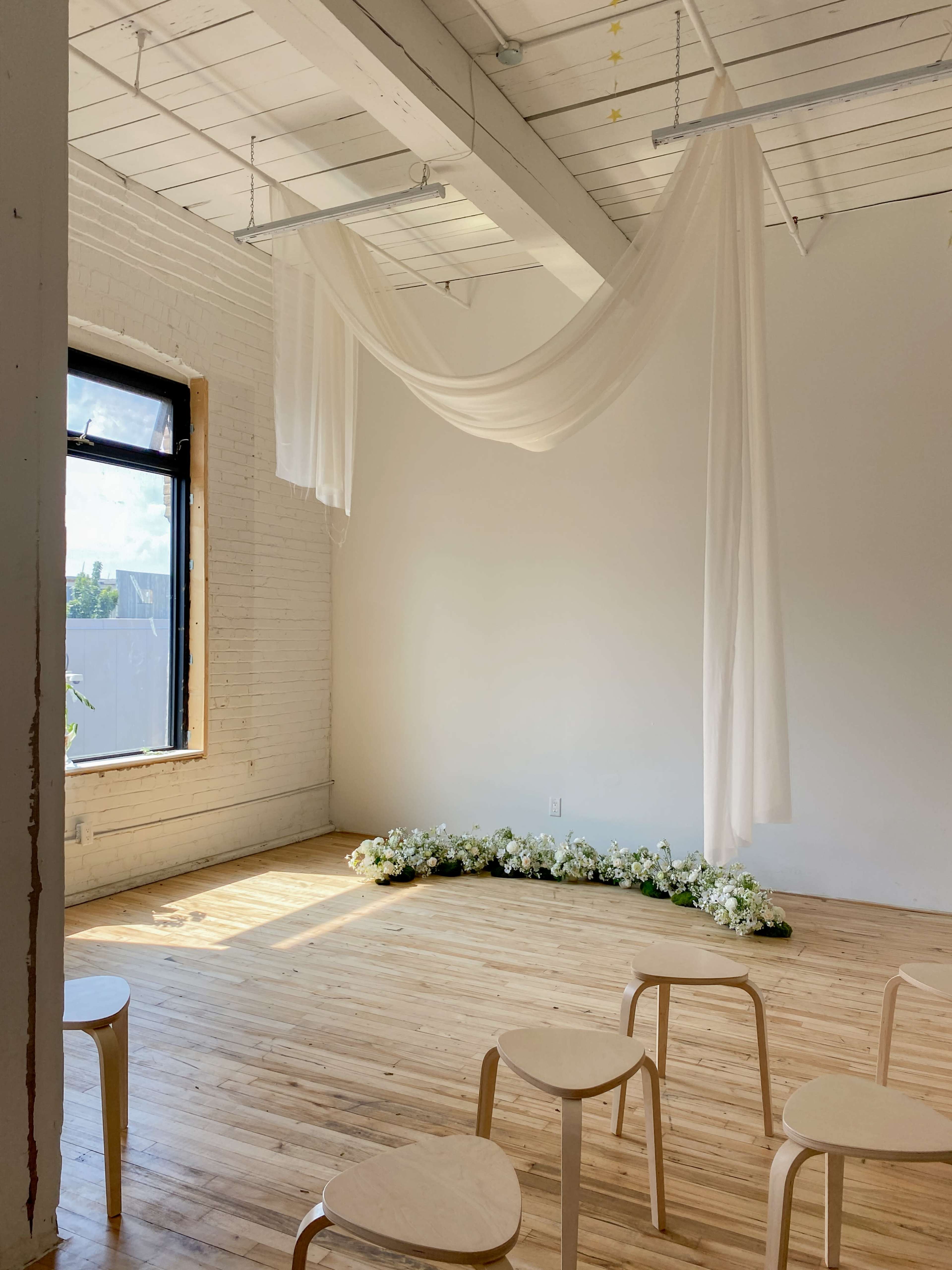 The image shows a bright, minimalist room with wooden flooring, light fabric draped from the ceiling, and a semicircle of artificial flowers on the floor, with several wooden stools arranged nearby.