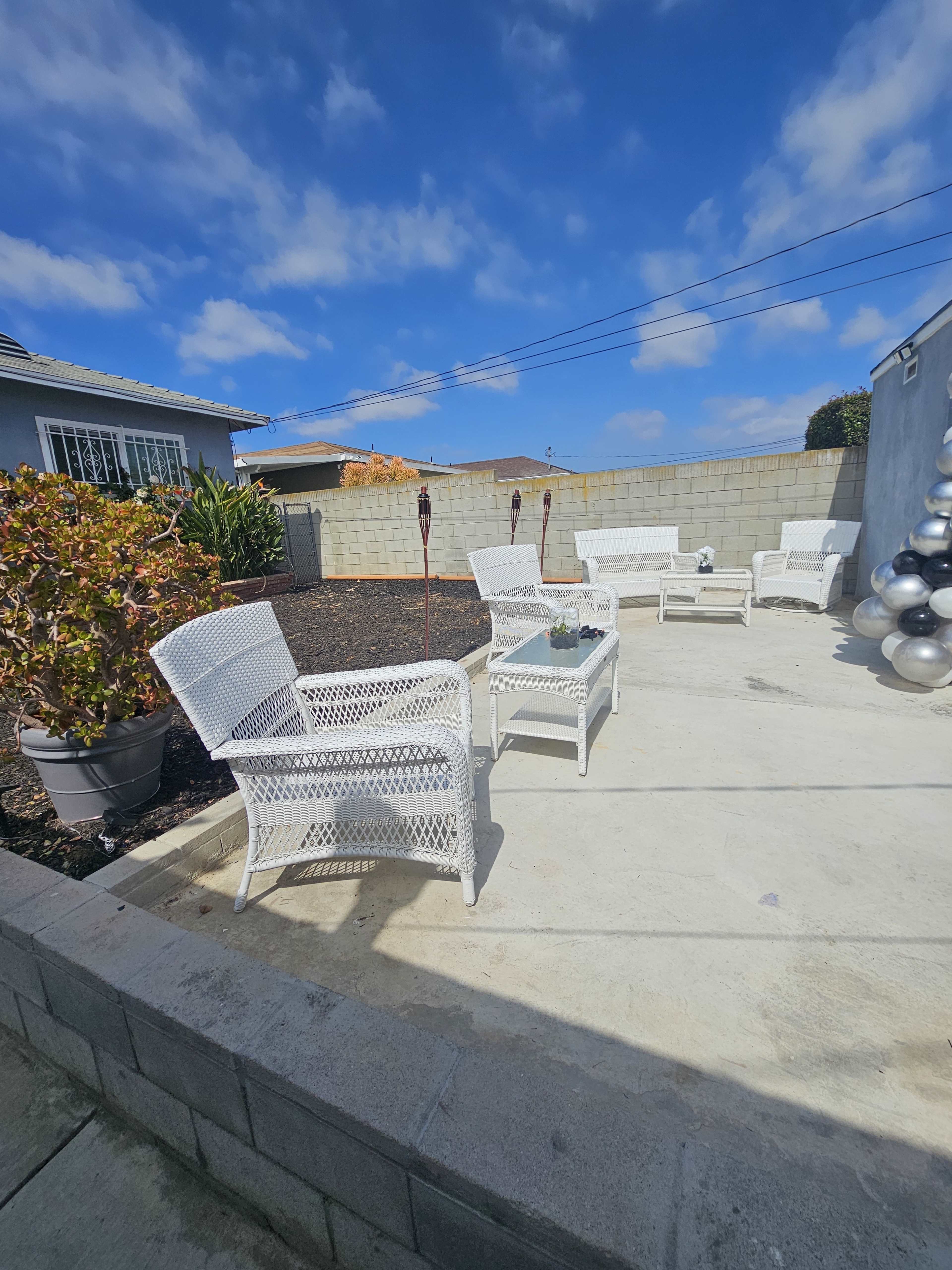 A patio area features white wicker chairs and a small potted plant, surrounded by a low wall and blue sky with clouds.