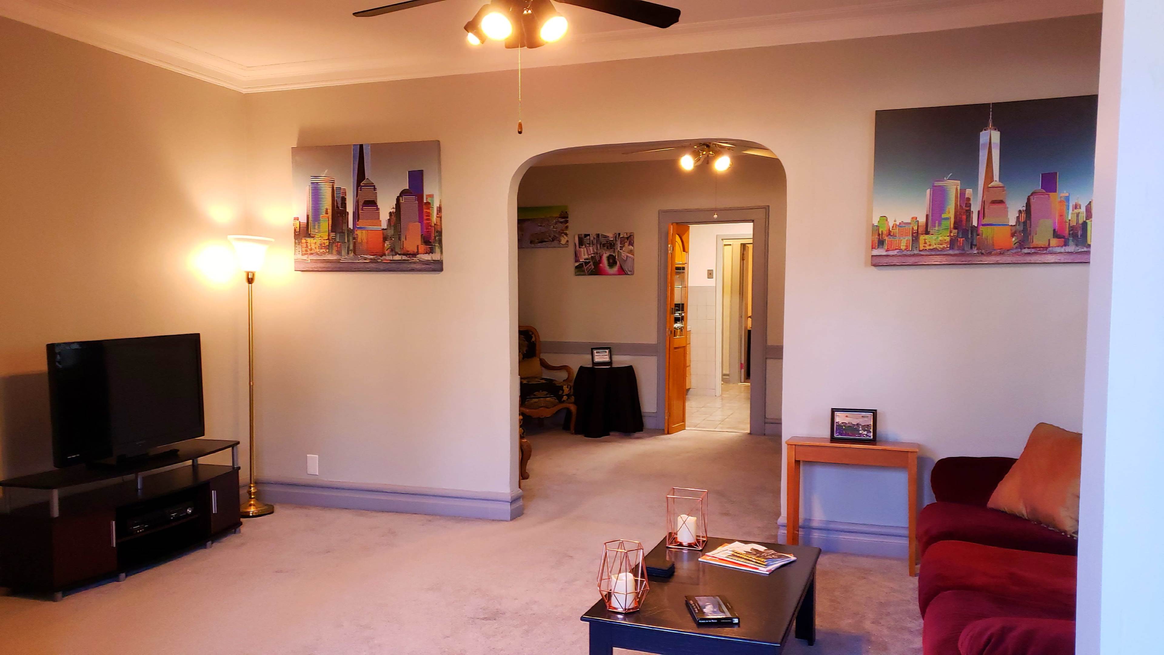 The living room features a television, a coffee table, a red couch, and two cityscape paintings on the walls.