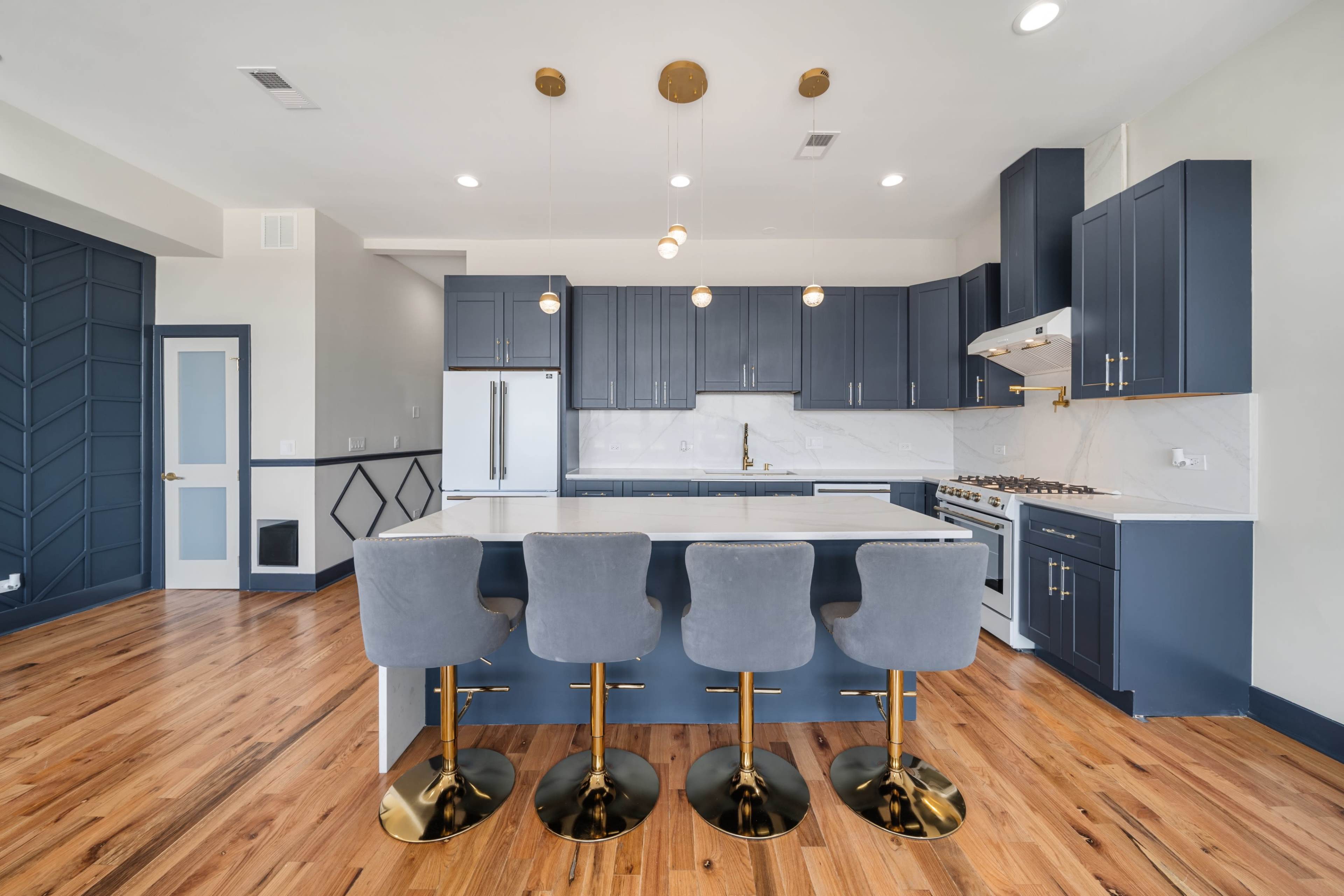 The image shows a modern kitchen featuring blue cabinets, a marble countertop, gold bar stools, and pendant lighting.
