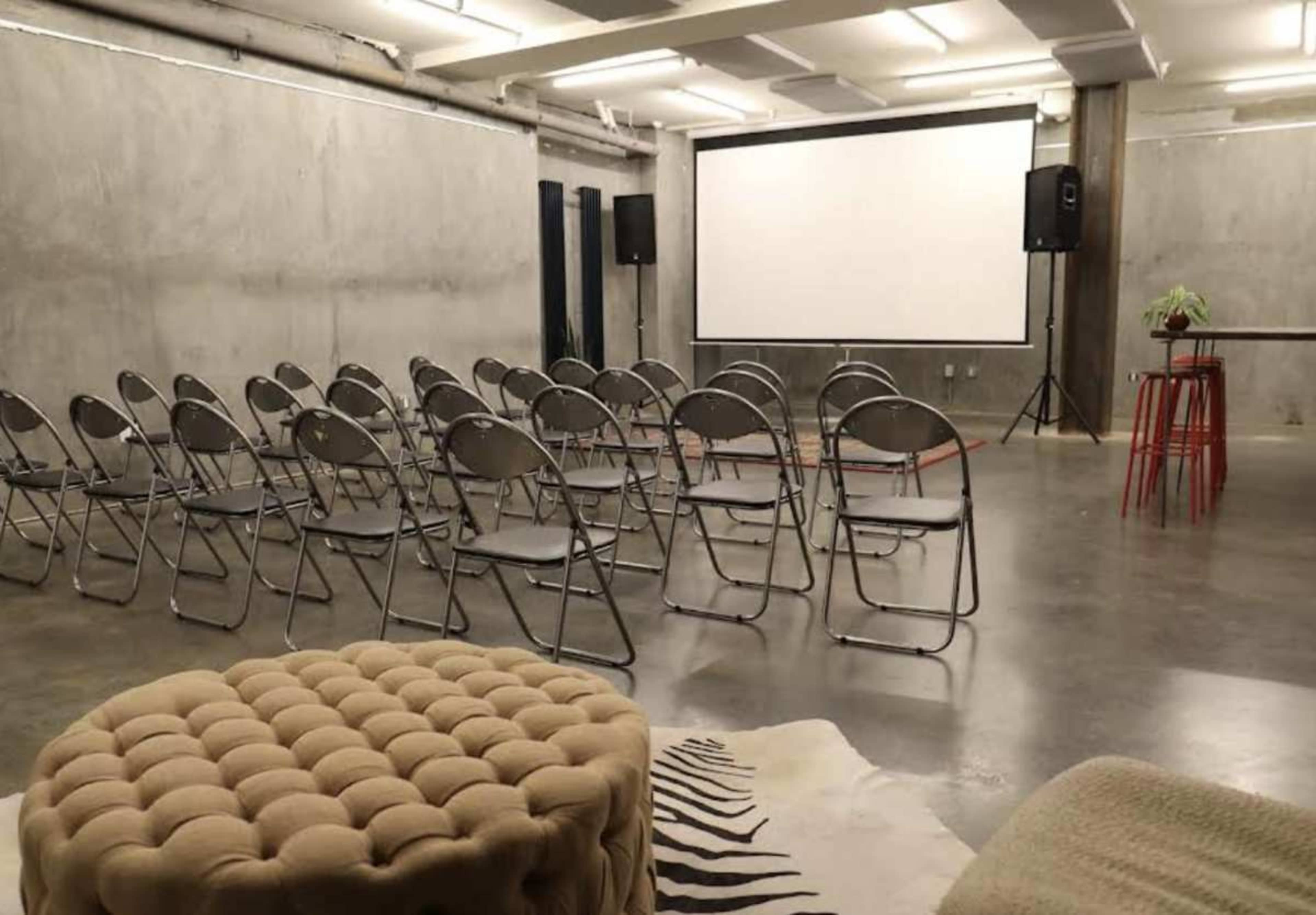 The image shows a minimalist event space with gray concrete walls and a projector screen at the front, arranged with rows of metal folding chairs and a small table nearby.