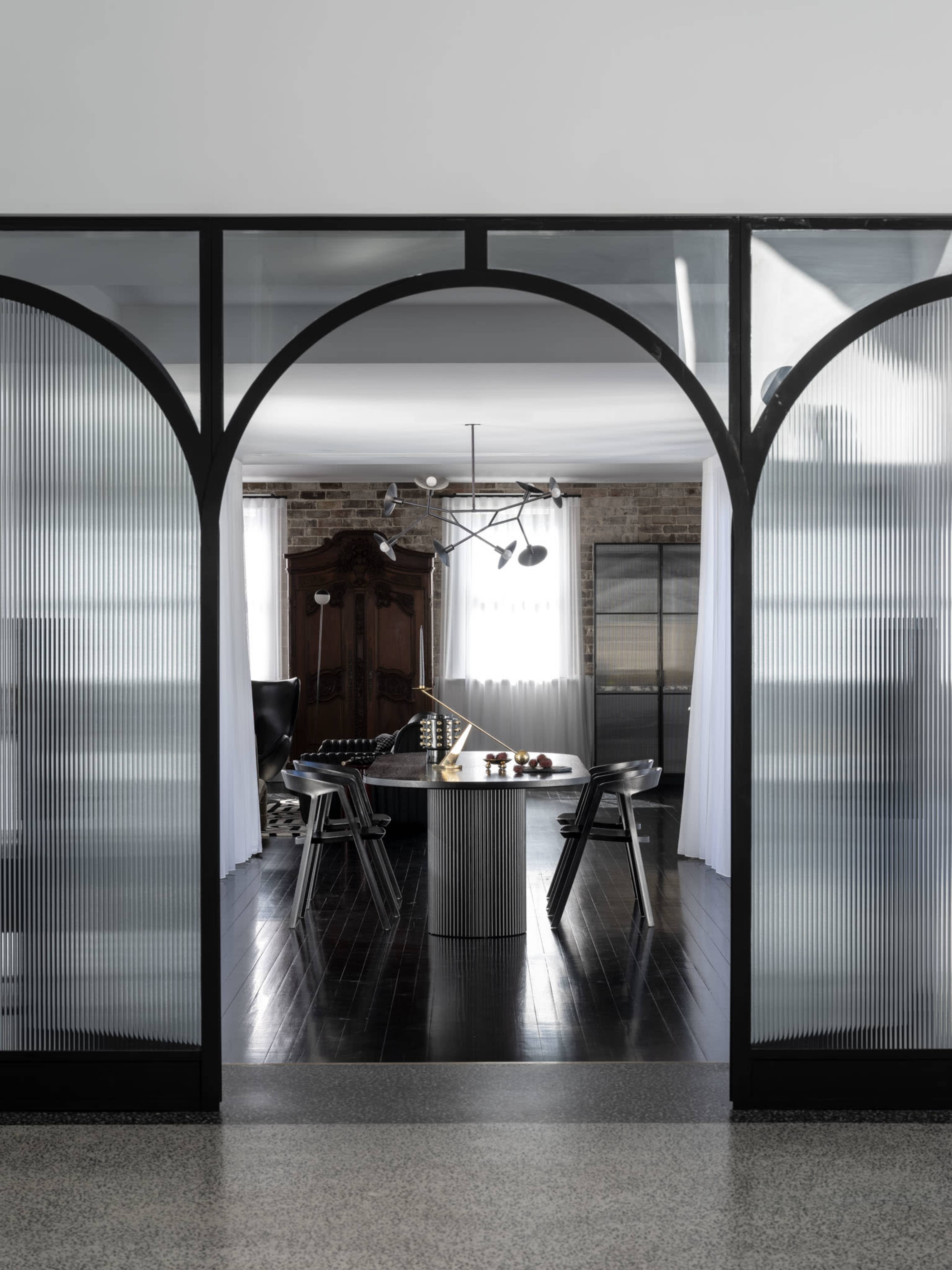 The image shows a modern interior featuring a dining area with chairs and a central table, framed by large, arched glass doors.