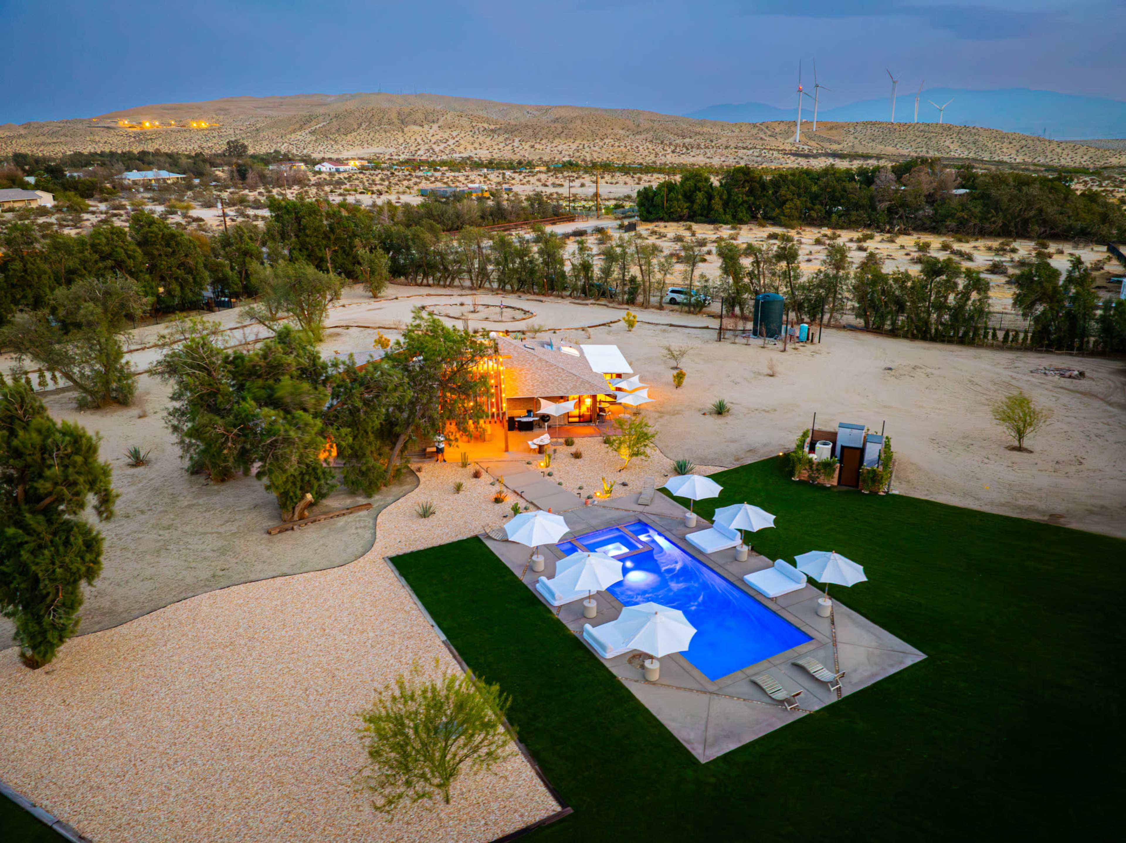 A modern desert villa with a pool, surrounded by palm trees and outdoor seating, sits against a backdrop of mountains and wind turbines.