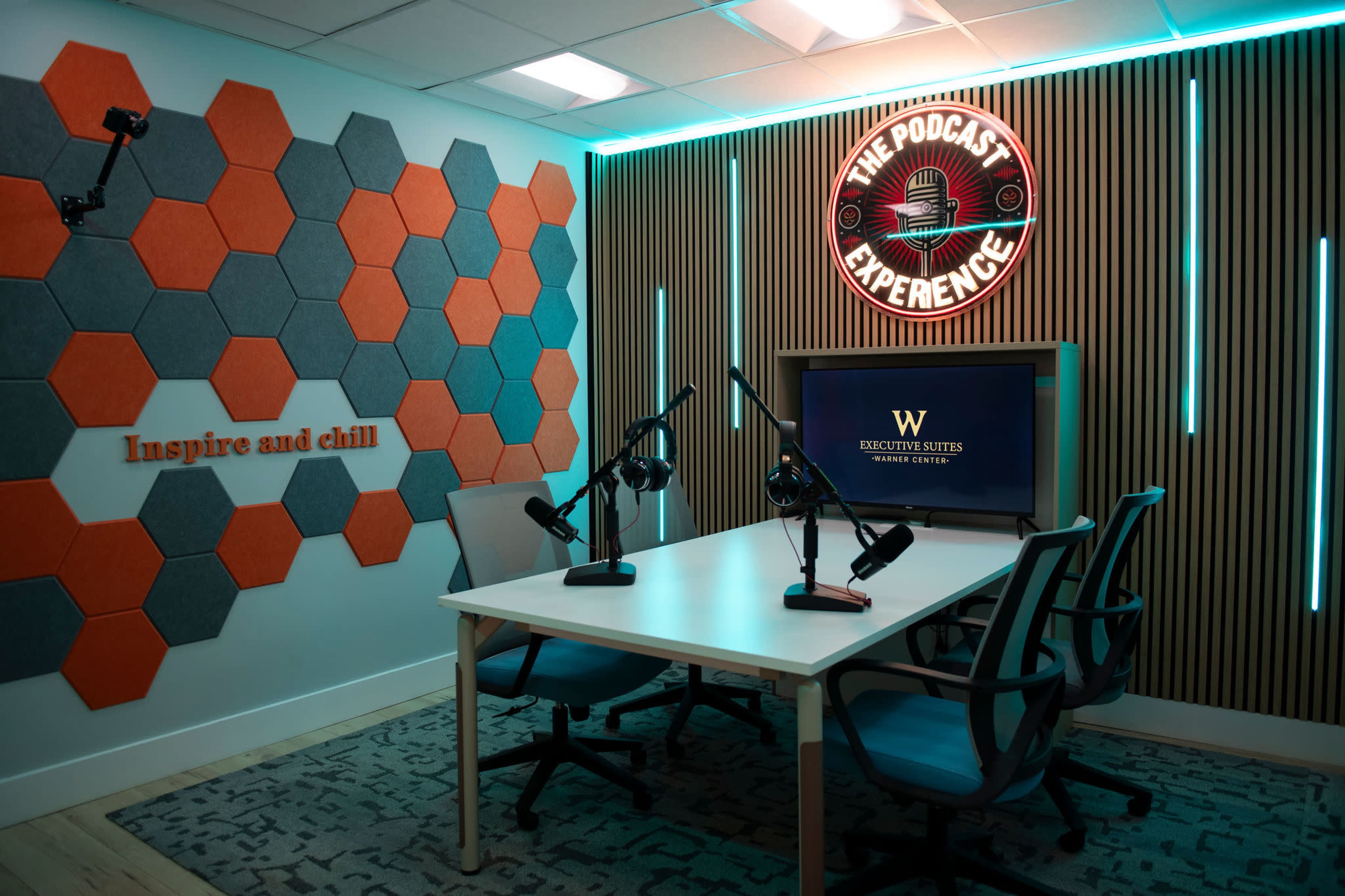 The image shows a podcast studio featuring a table with microphones, a screen displaying text, and colorful wall decor made of hexagonal panels.
