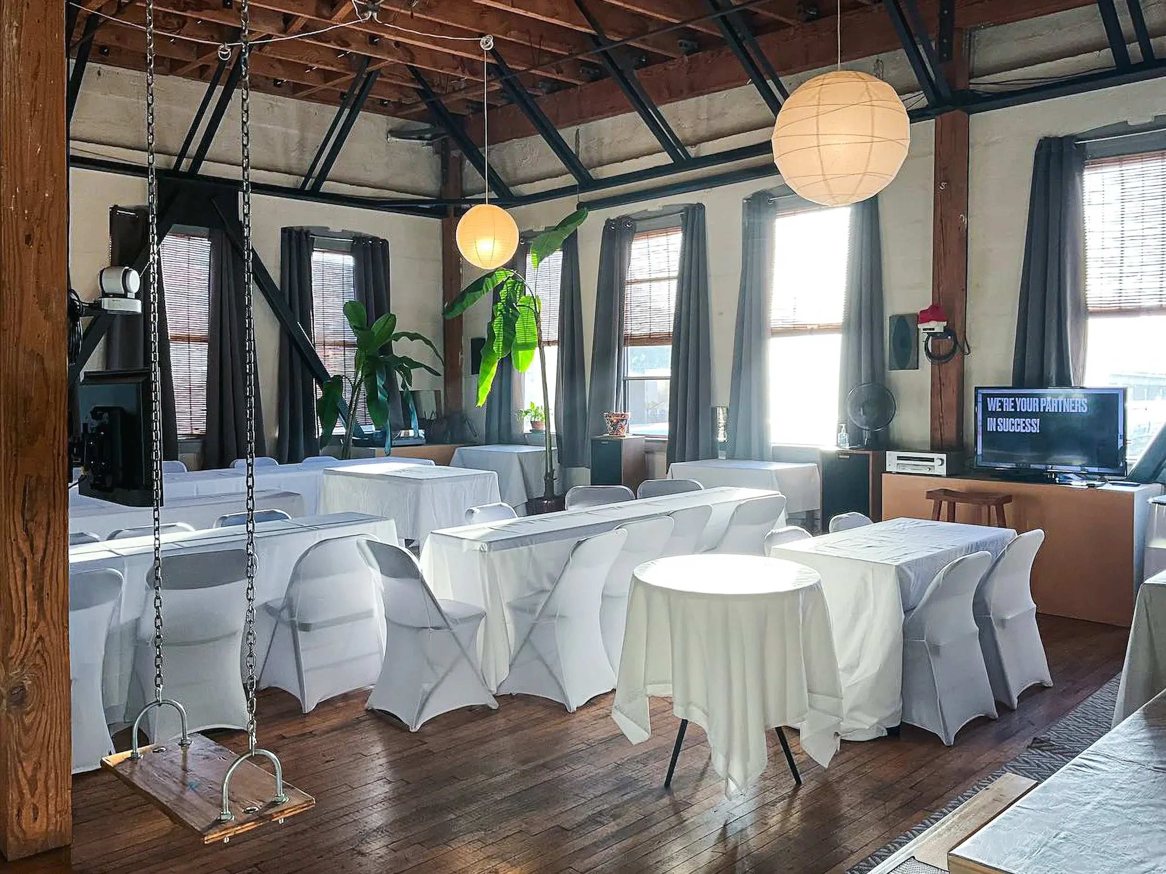 The image shows a spacious room with wooden beams, large windows, and tables covered with white cloths, set up for an event or meeting.