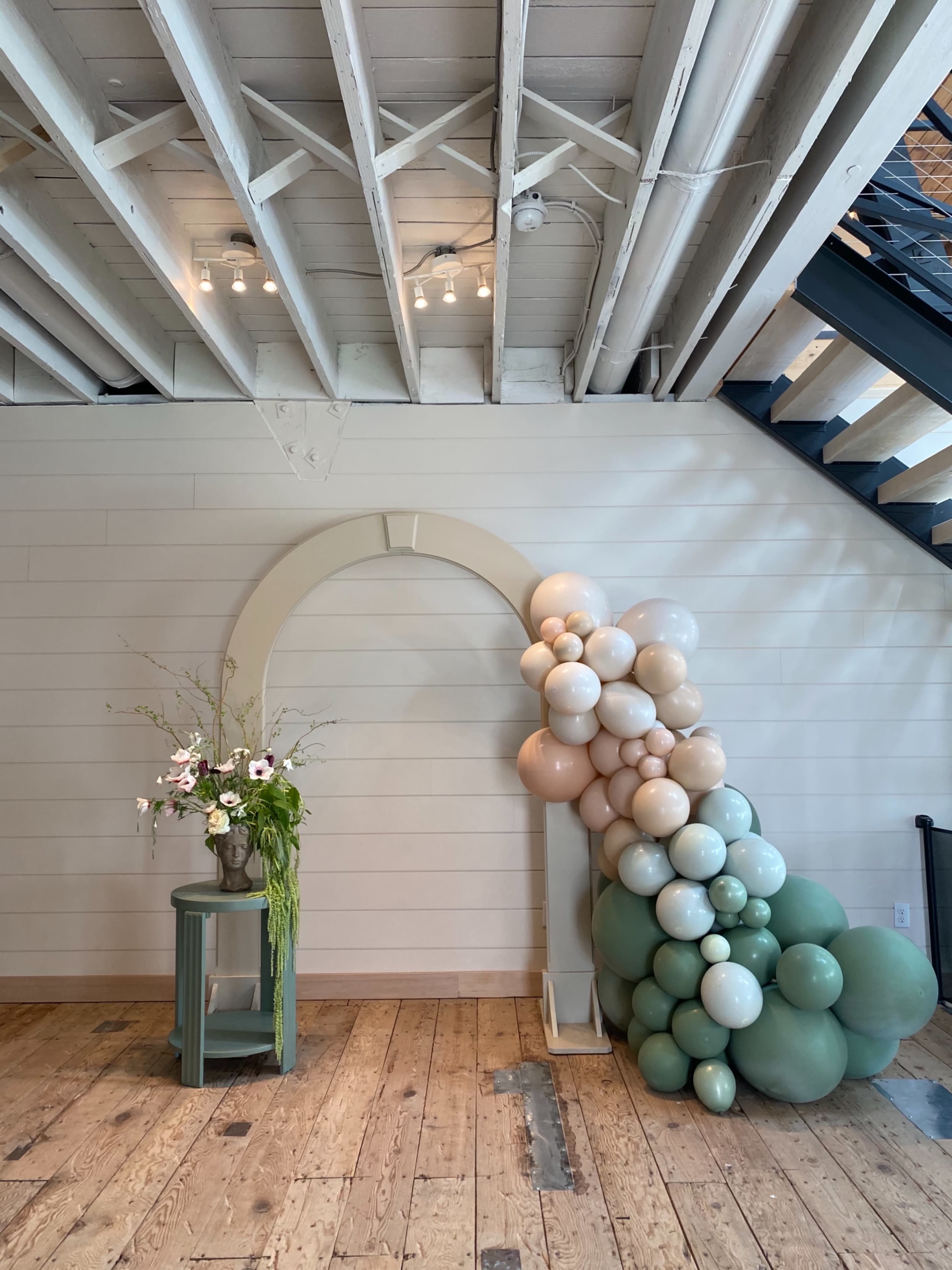 A decorative arch made of balloons in neutral tones stands next to a green side table with a floral arrangement, against a backdrop of wooden flooring and a staircase.