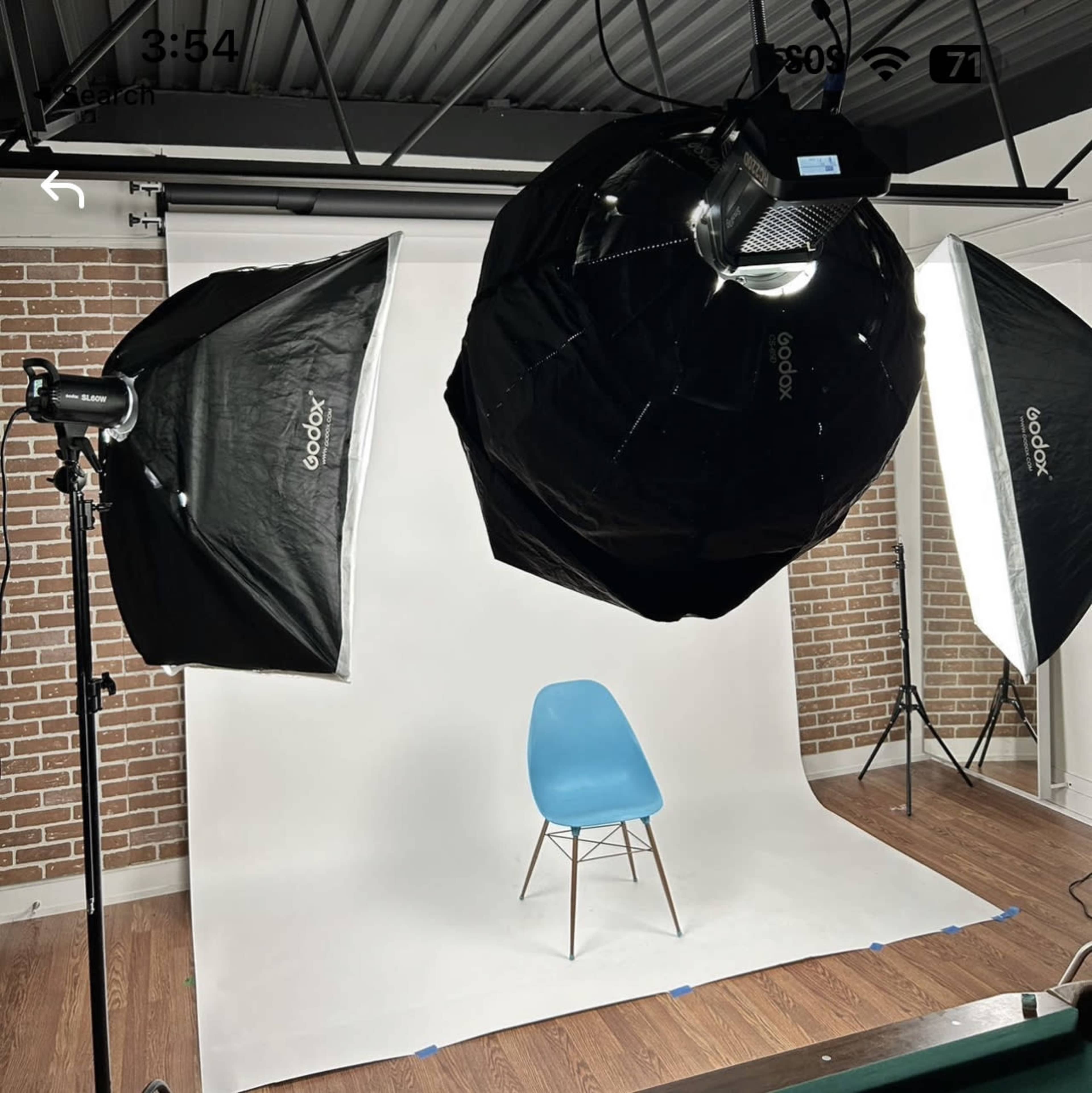 A photography studio setup with two softbox lights, a large round light overhead, and a single blue chair positioned on a white backdrop against a brick wall.