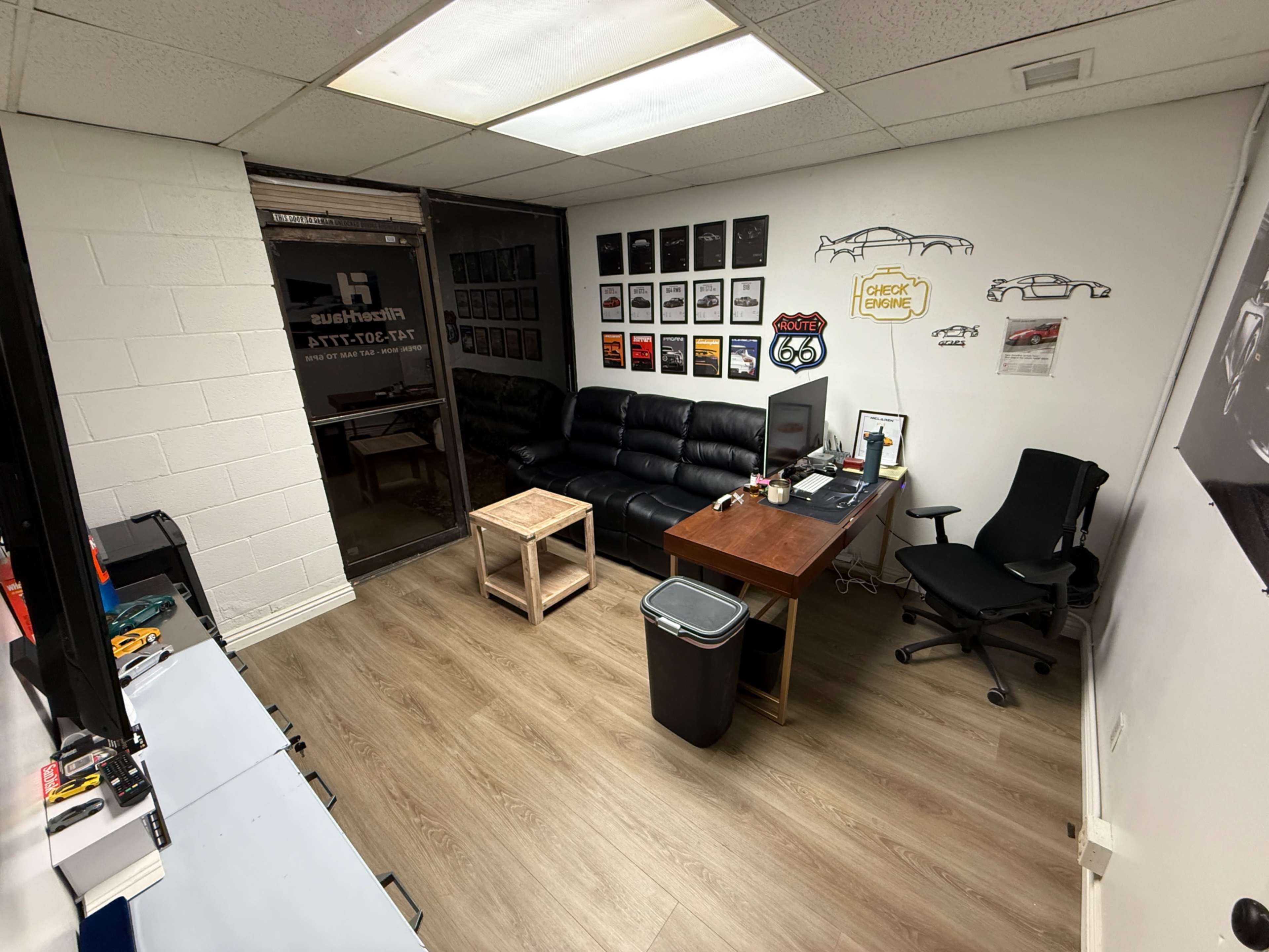 The image shows a small office space featuring a black leather couch, a wooden table, a desk with a computer and office chair, and several framed pictures on the walls.