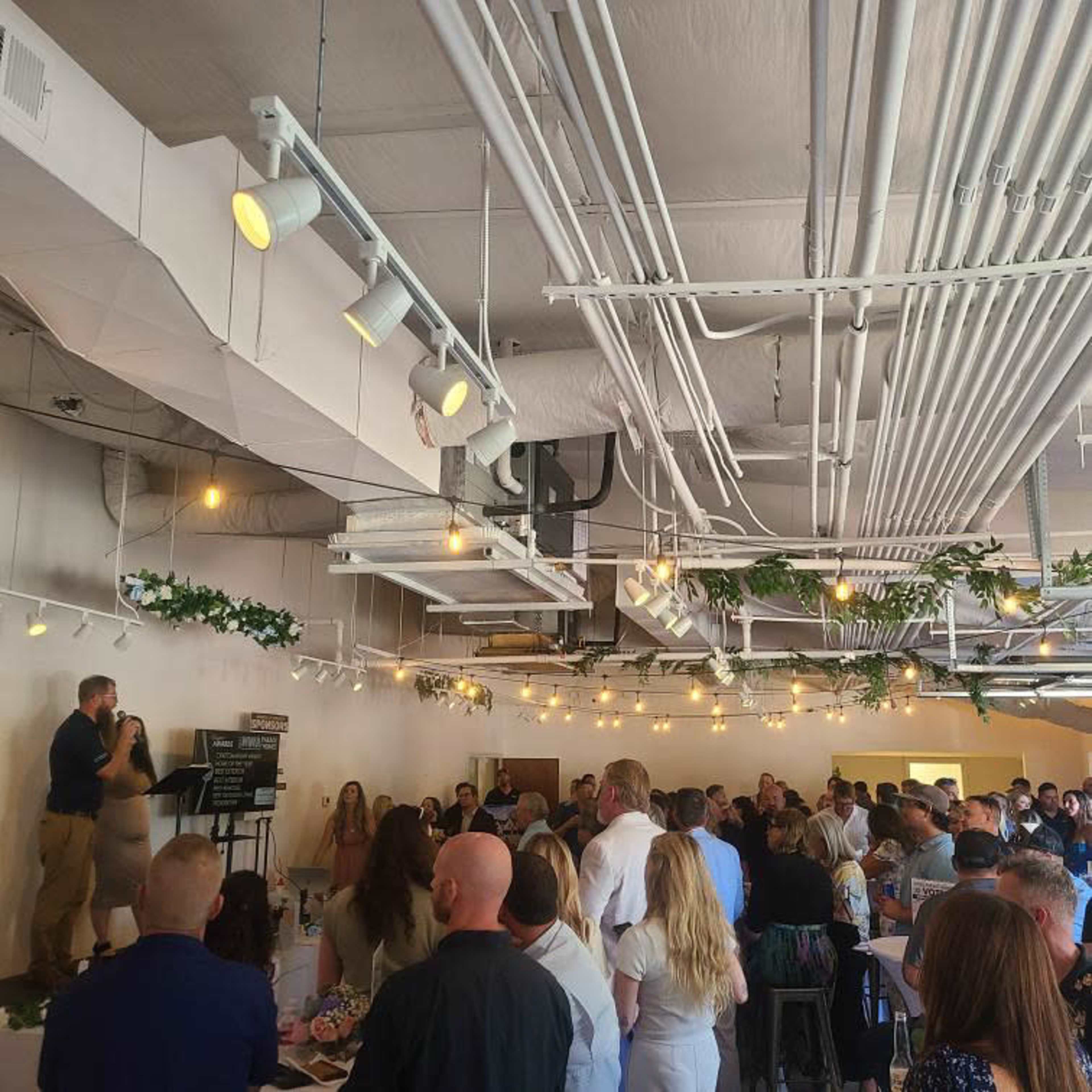 A crowd gathers in a well-lit venue with decorative lighting and plants, as two speakers address the audience from a small stage.
