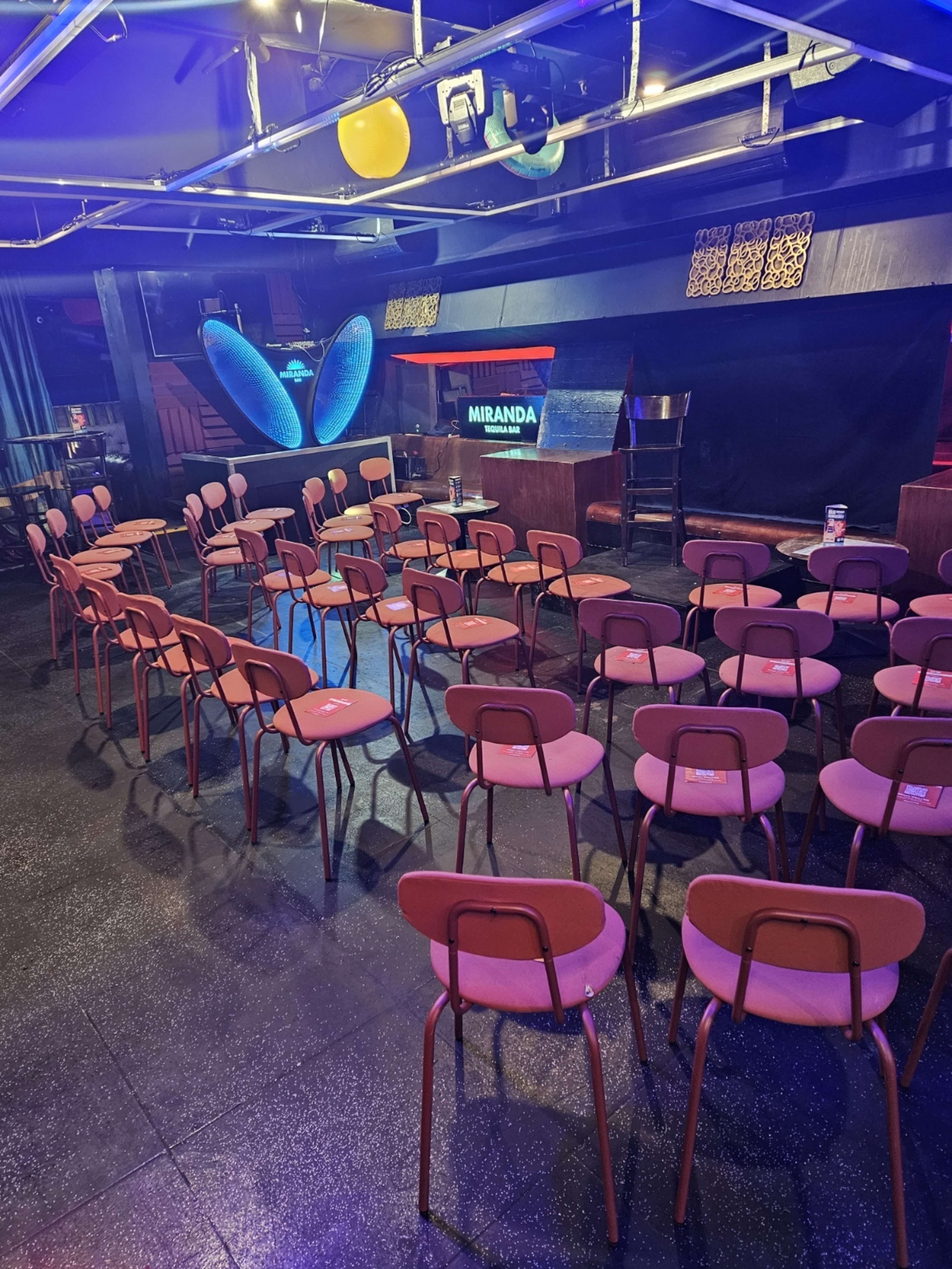 The image shows a stage setup with rows of empty pink chairs arranged in a venue.