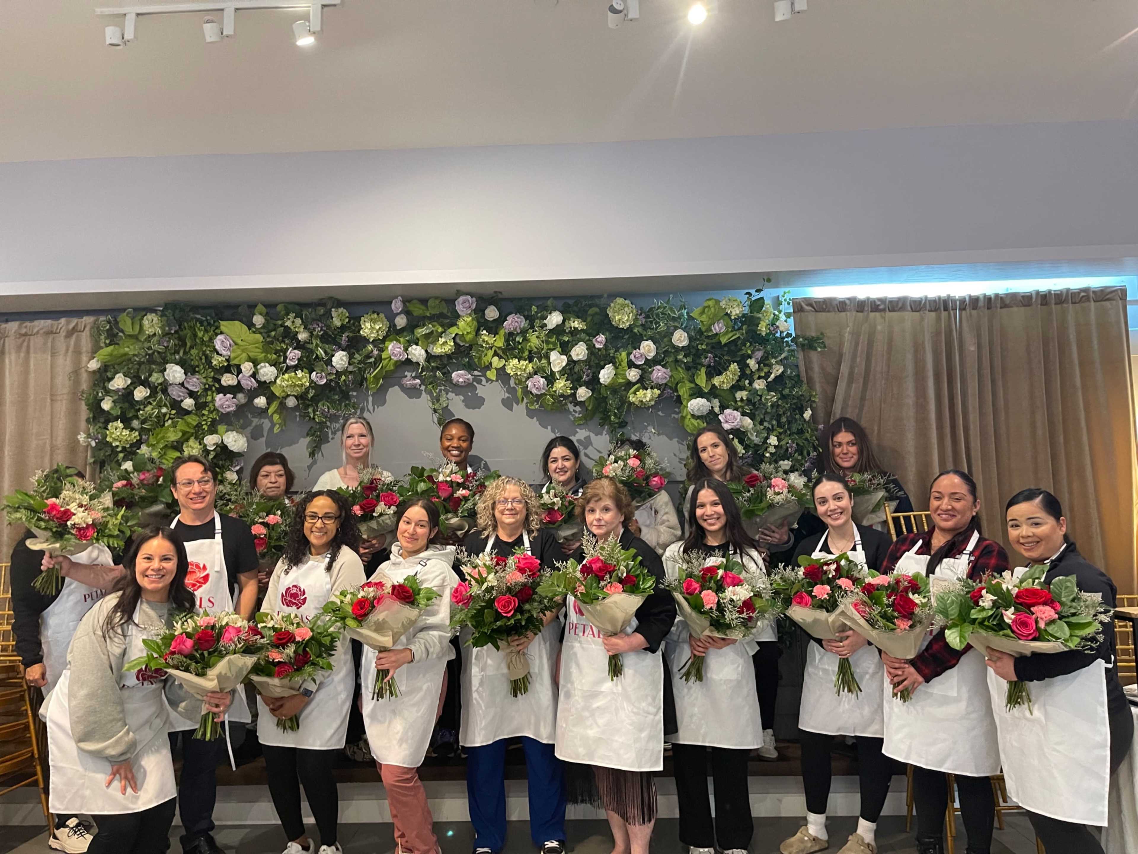 A group of twenty people stands together in front of a floral backdrop, each holding colorful bouquets of flowers.