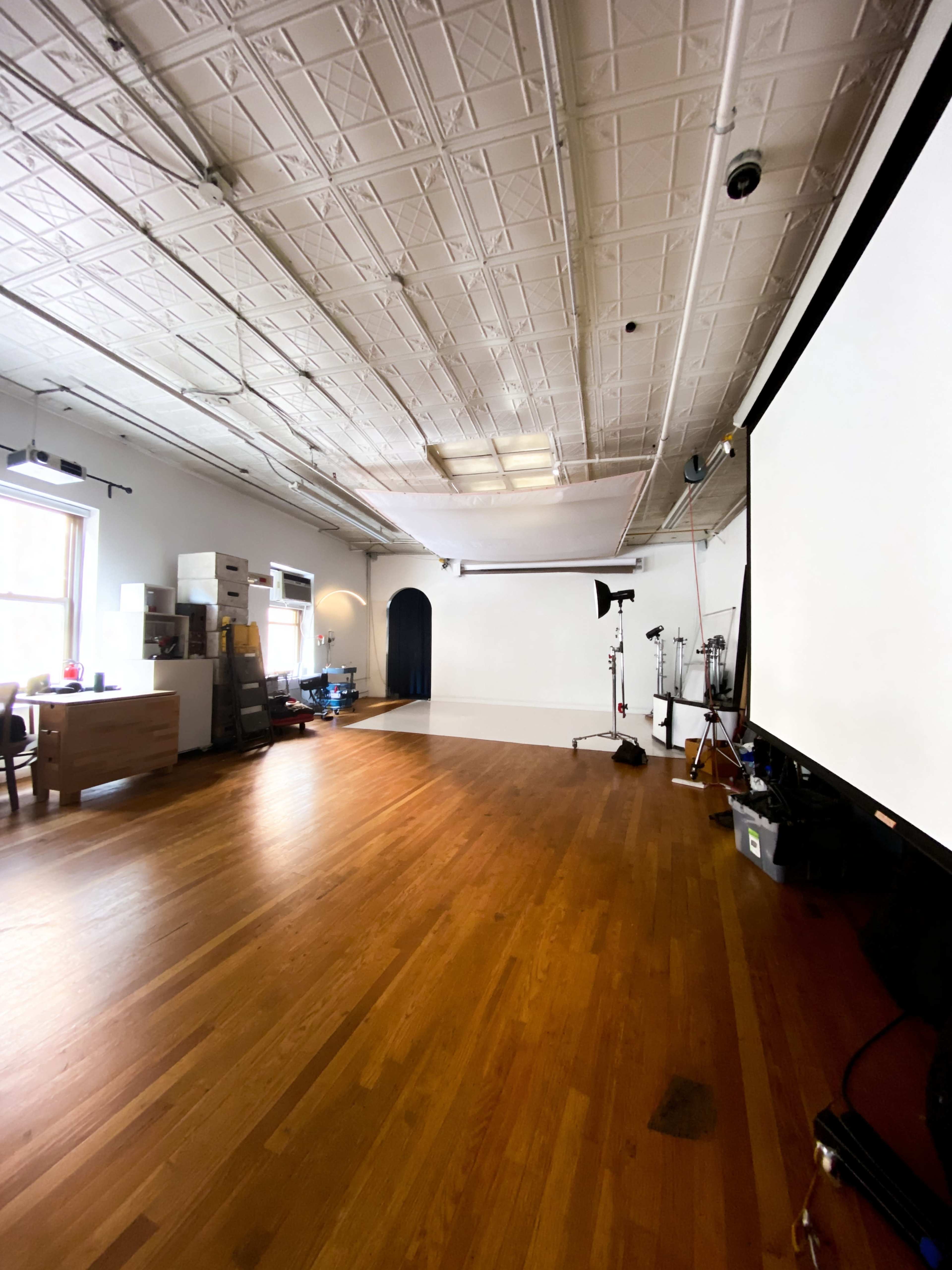 The image shows a spacious, empty studio with wooden flooring, large windows, and a blank white wall at the front, along with some equipment and storage in the back.