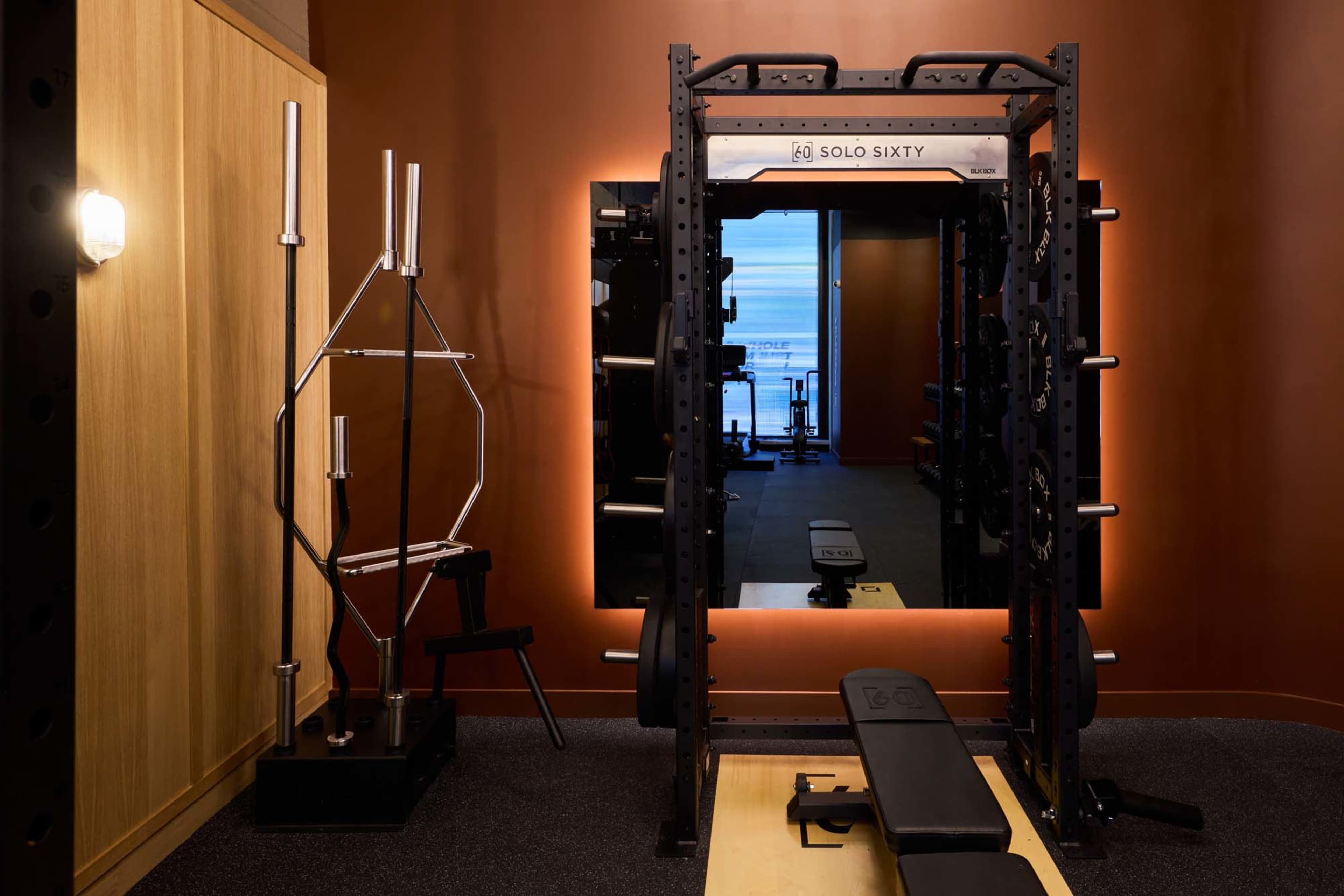 The image shows a gym area featuring a power rack, a weight bench, and a mirrored wall, all set against a warm-toned backdrop.