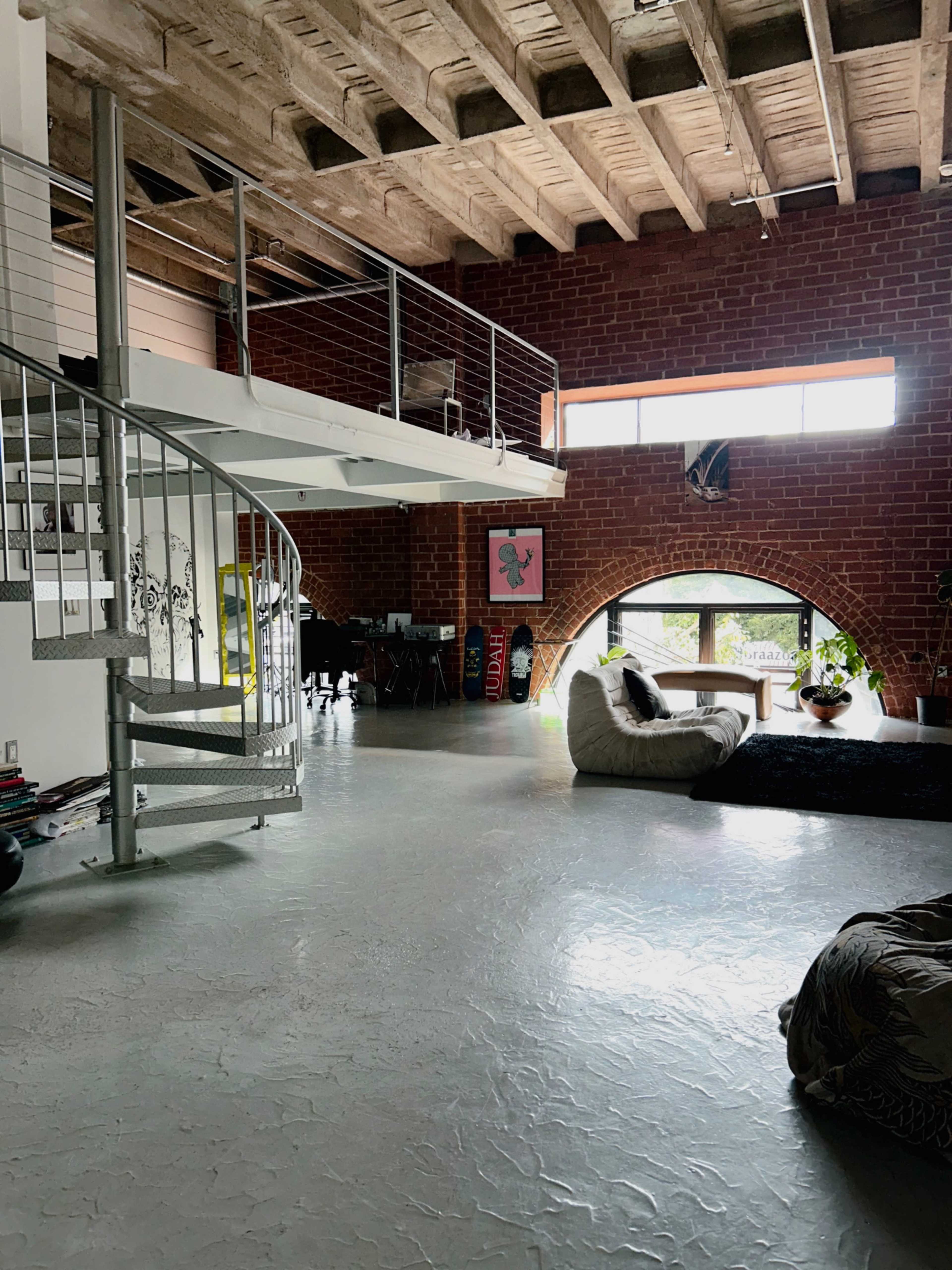 The image shows a spacious industrial-style loft with exposed brick walls, a spiral staircase leading to a mezzanine, and large windows allowing natural light to fill the area.