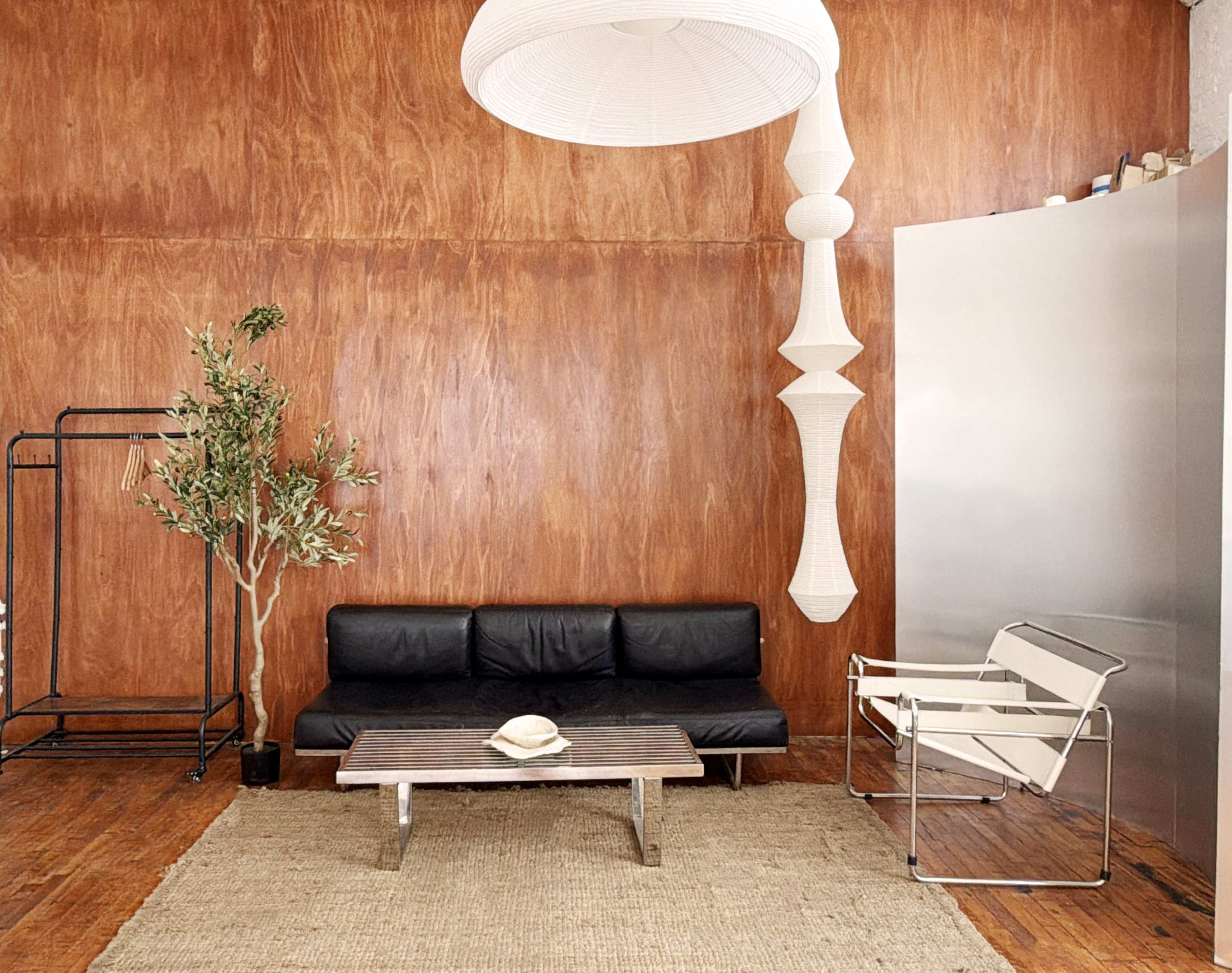 The image shows a minimalist living space with a black couch, a wooden coffee table, a plant, and a large metallic cabinet against a wooden wall.