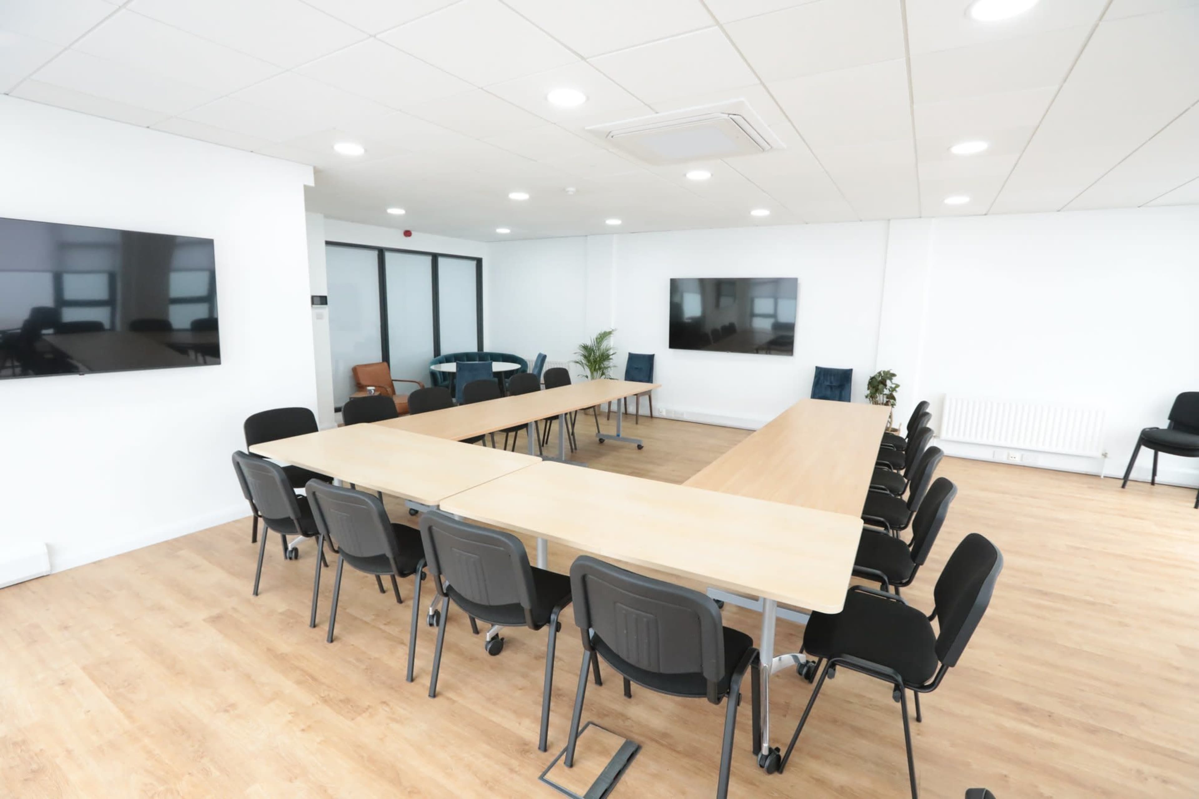A large conference room features a long rectangular table surrounded by black chairs, with two screens mounted on the white walls.