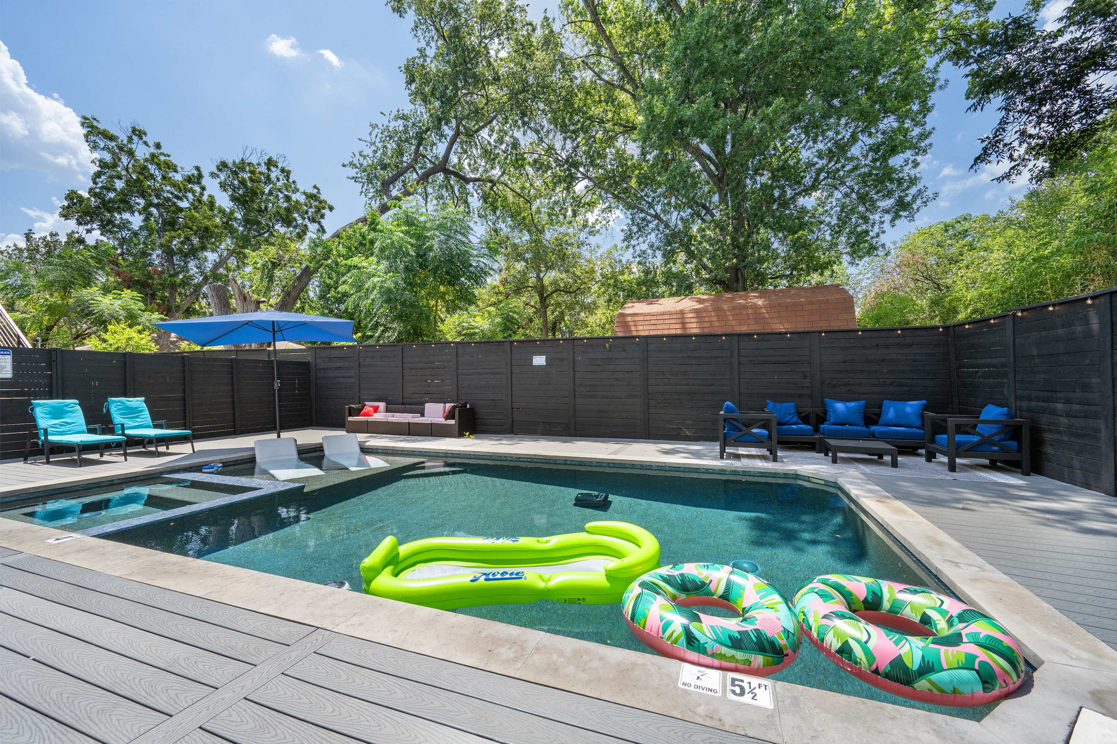 The image shows a backyard pool area with a clear swimming pool, two colorful pool floats, lounge chairs, and shaded seating, surrounded by a wooden fence and trees.