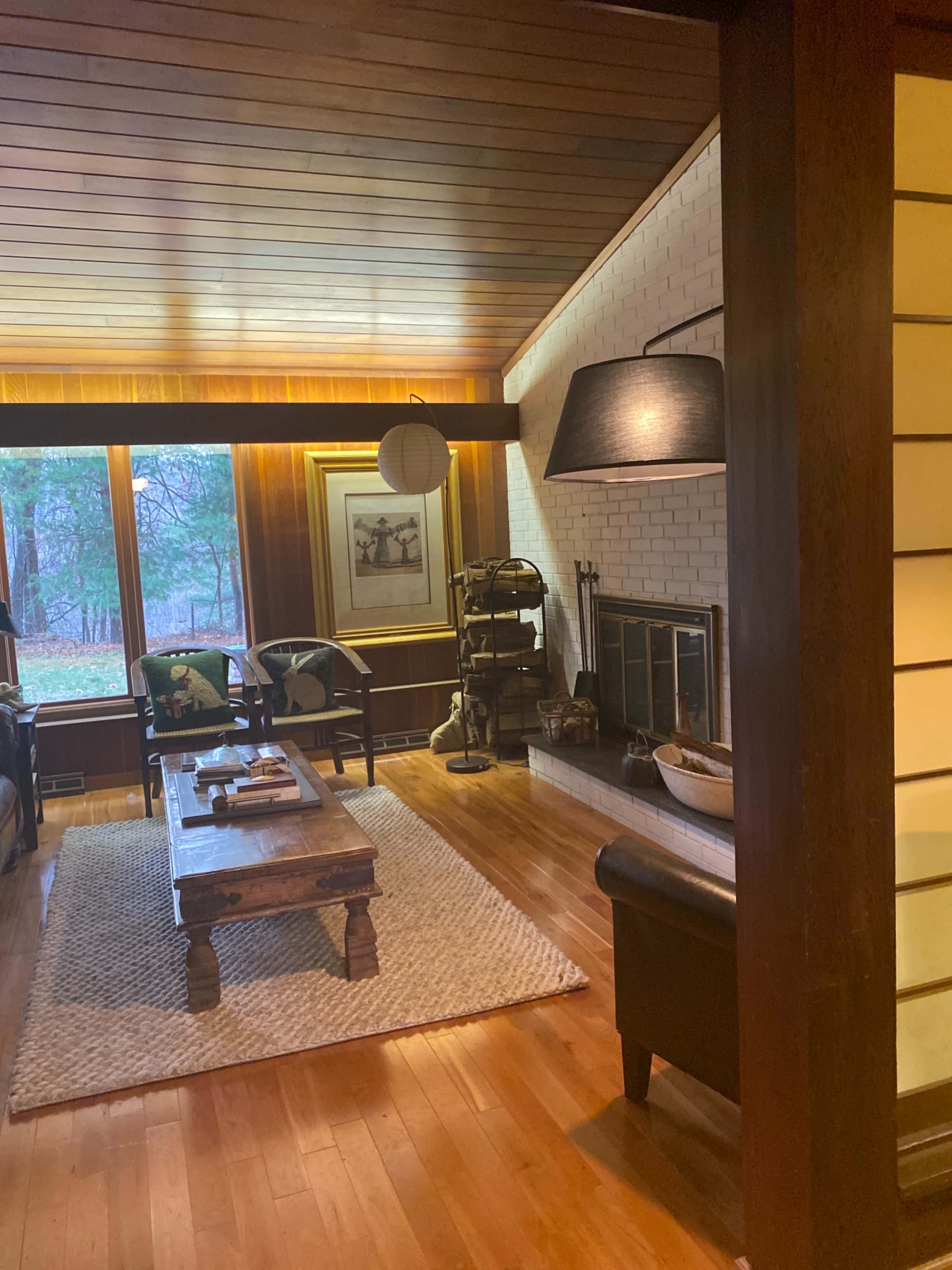 The image shows a cozy living room featuring a wooden ceiling, a fireplace, and two chairs flanking a coffee table on a woven rug.
