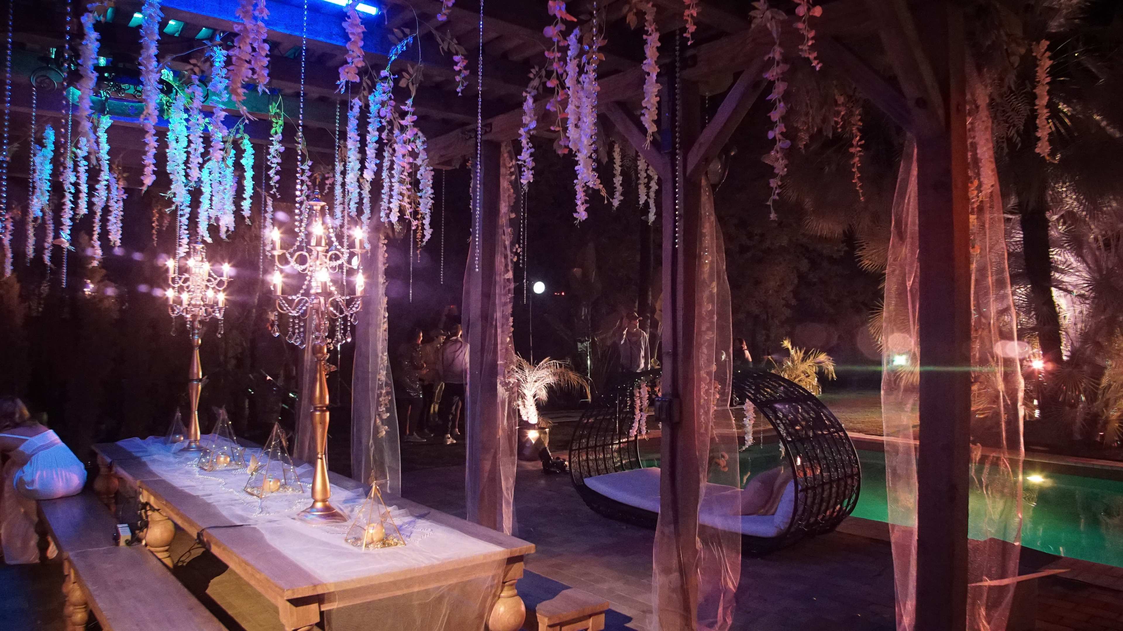 A beautifully decorated outdoor space featuring a long dining table, elegant chandeliers, and a pool surrounded by tropical foliage.