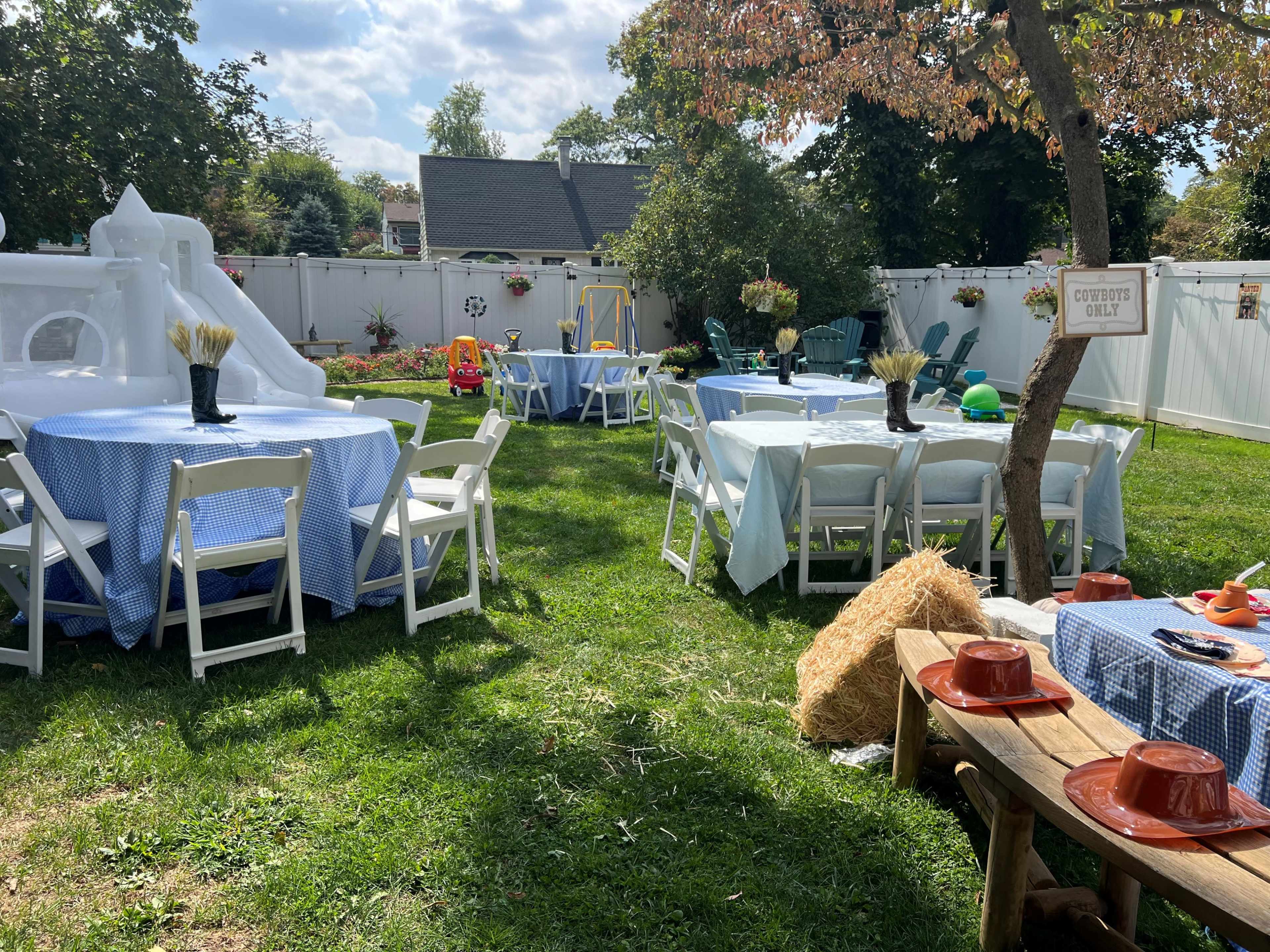 The image shows an outdoor space set up for a gathering, featuring several tables covered in blue and white tablecloths, a play structure in the background, and decorations scattered around the lawn.