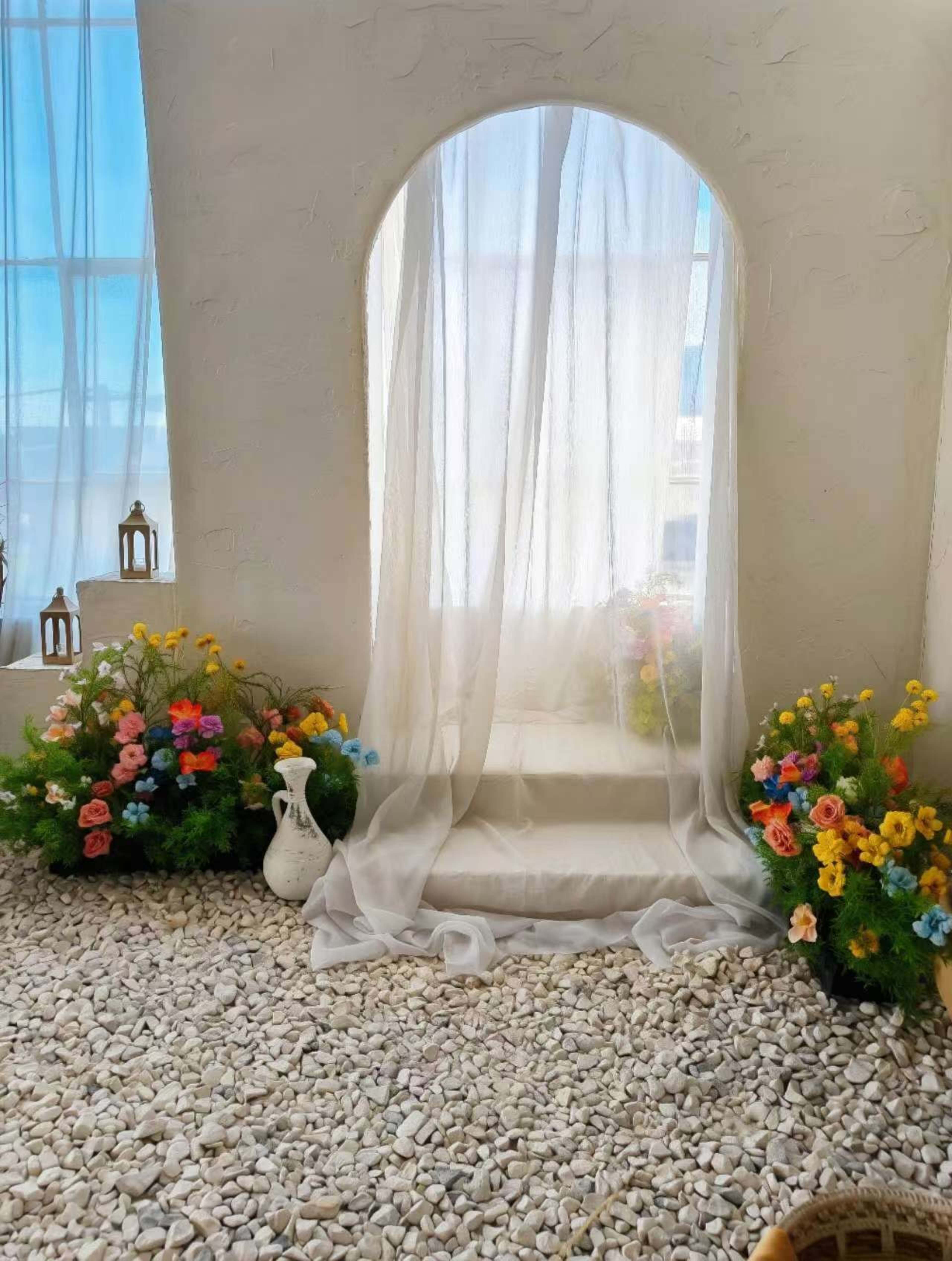 A sheer white curtain drapes over an arched doorway, flanked by flower arrangements and set on a pebbled surface.