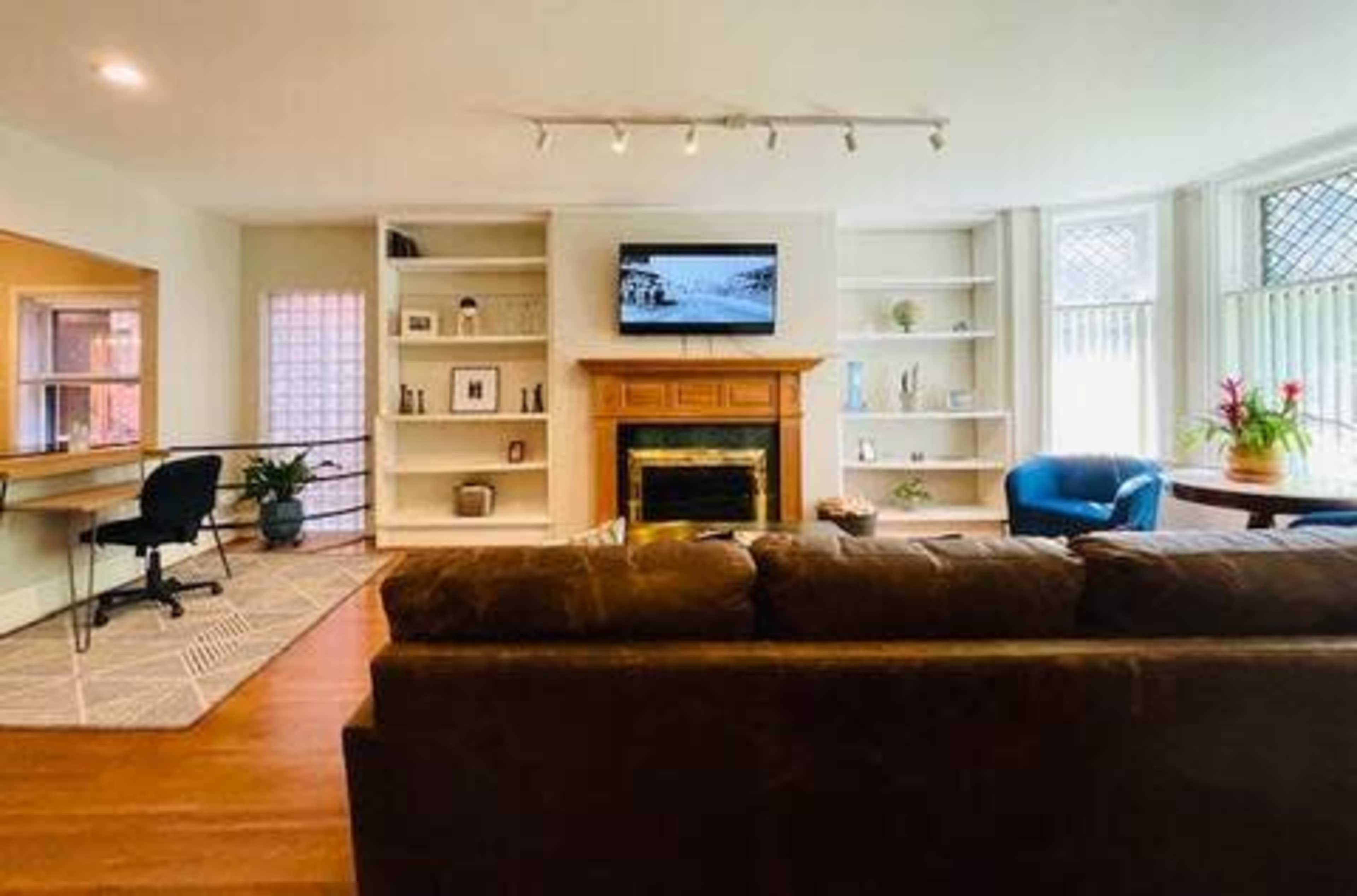 A spacious living room features a brown couch, a fireplace with a television above it, built-in shelves, and a desk area with a chair.