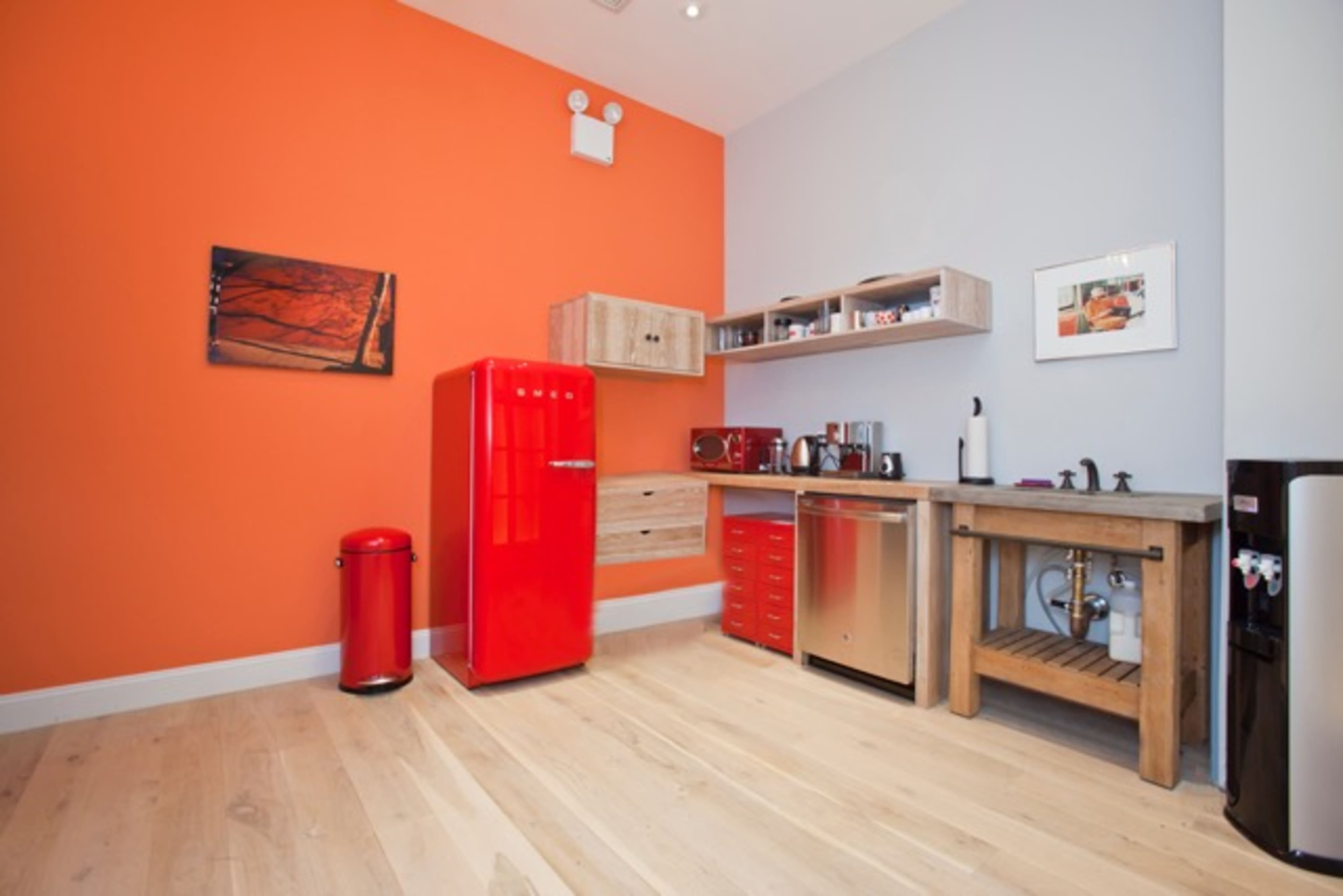A kitchen with an orange accent wall, featuring a red refrigerator, wooden shelving, and a mix of appliances on a wooden floor.