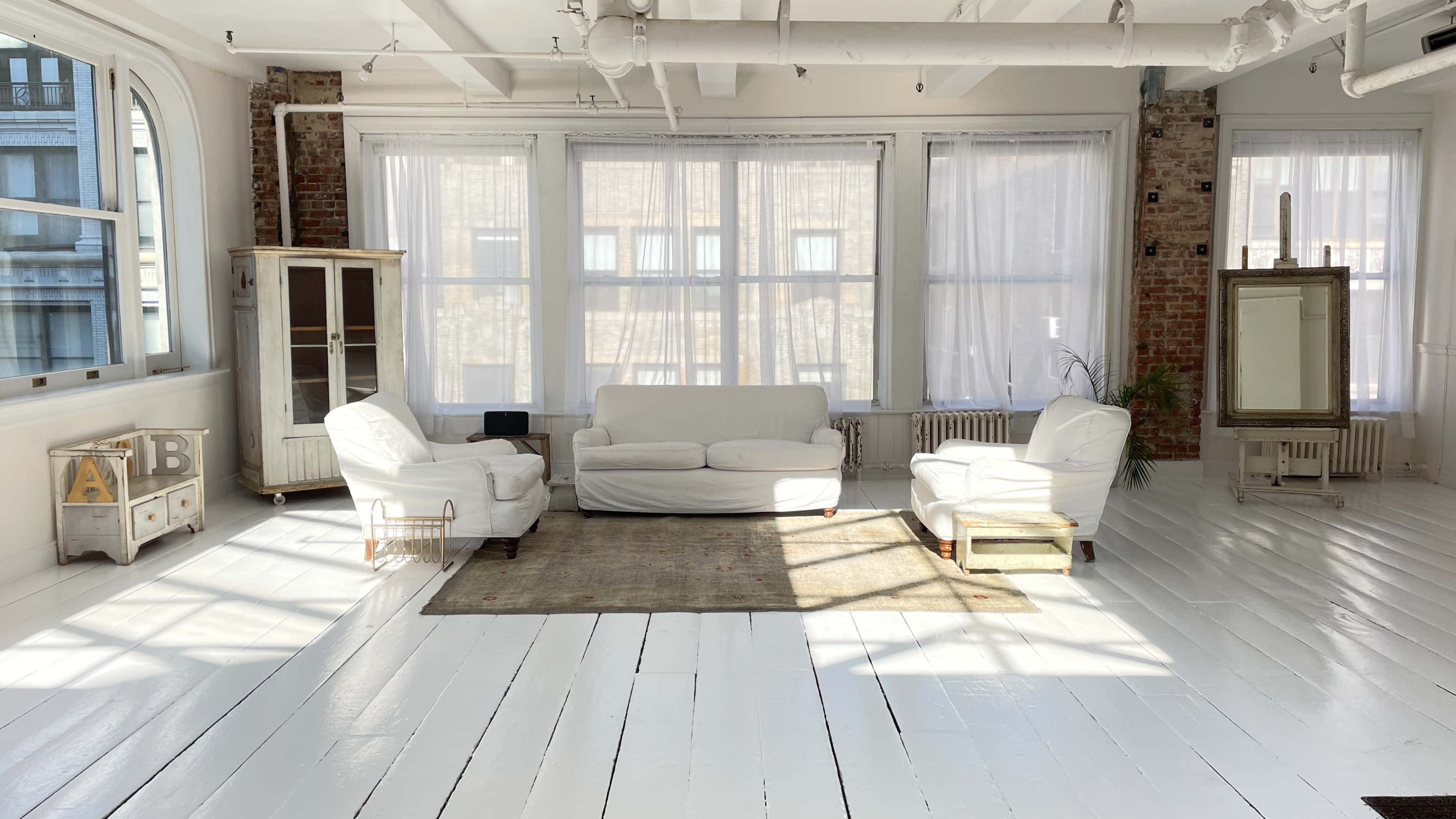 The image shows a bright, spacious room with white wooden floors, featuring two white armchairs, a white couch, a rug, and large windows letting in natural light.