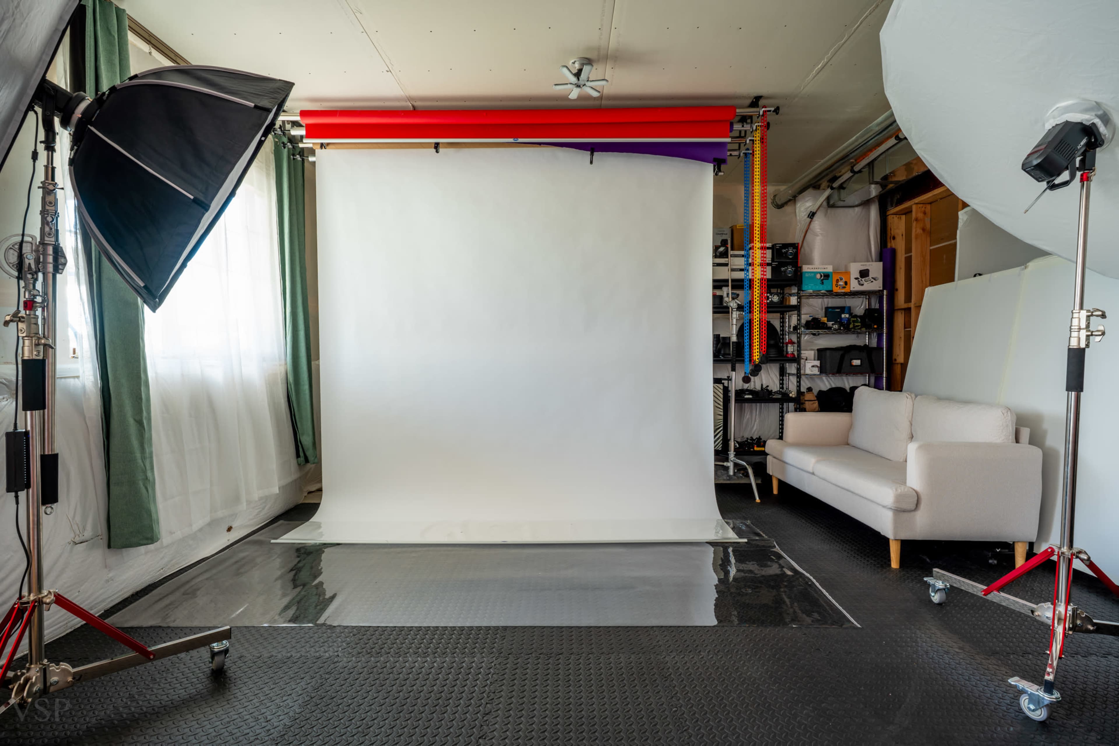 The image shows a photography studio with a white backdrop, softbox lighting, and a beige sofa against a wall with equipment and props in the background.