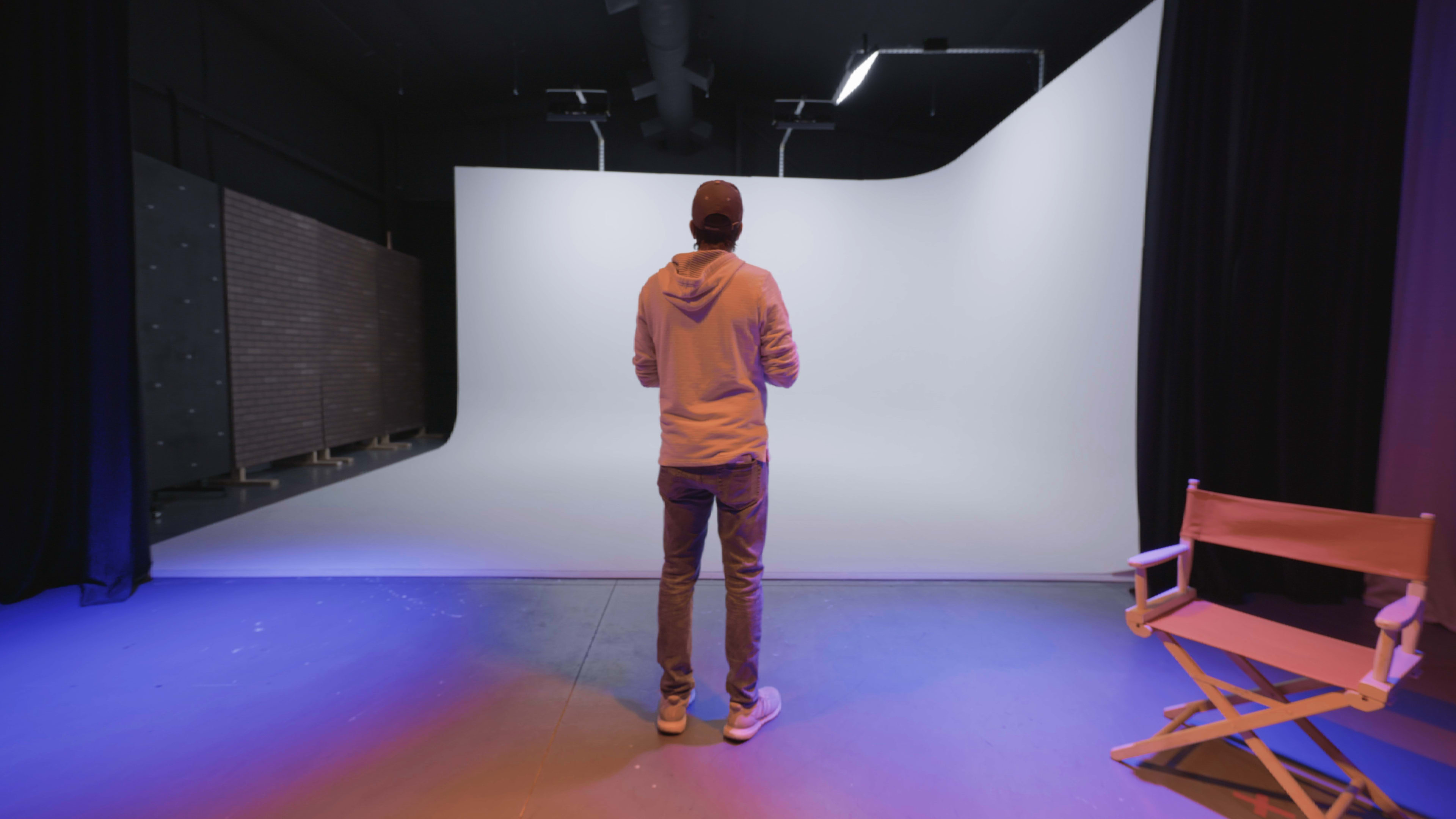 A person stands in front of a blank white backdrop in a studio, with a director's chair visible to the side.