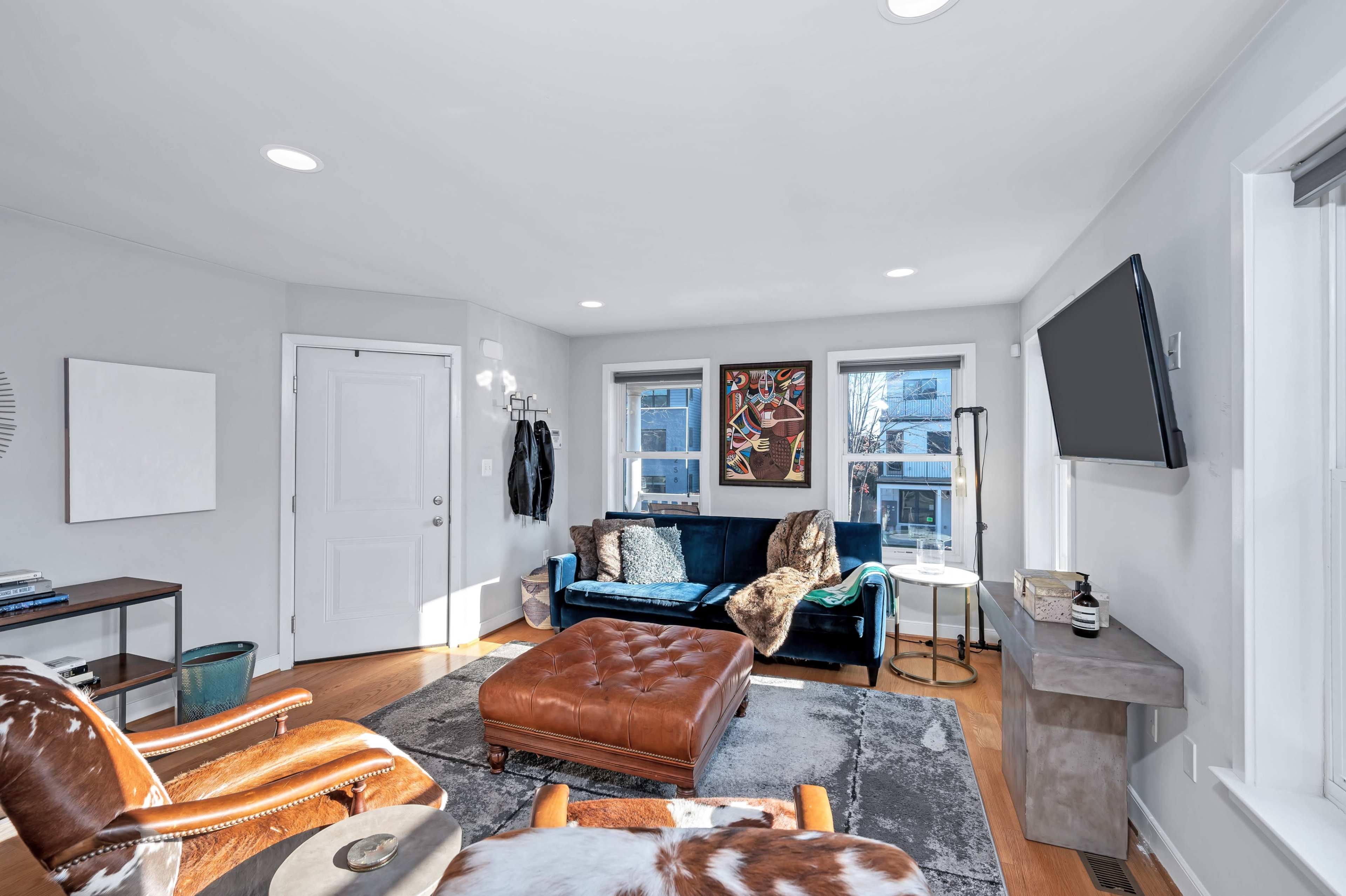 A modern living room featuring a blue sofa, a leather chair, an ottoman, and large windows allowing natural light.