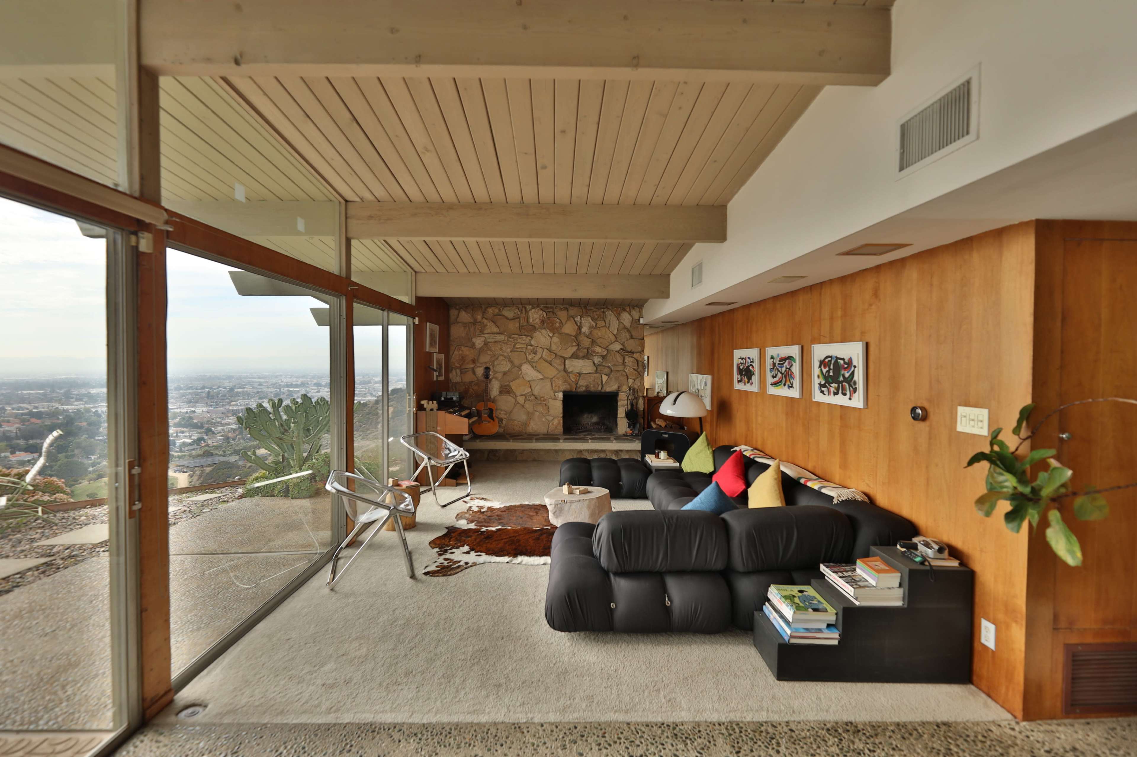 The image shows a modern living room with large glass windows, a stone fireplace, and a mix of furniture including a black sofa, a chair, and a decorative area rug.